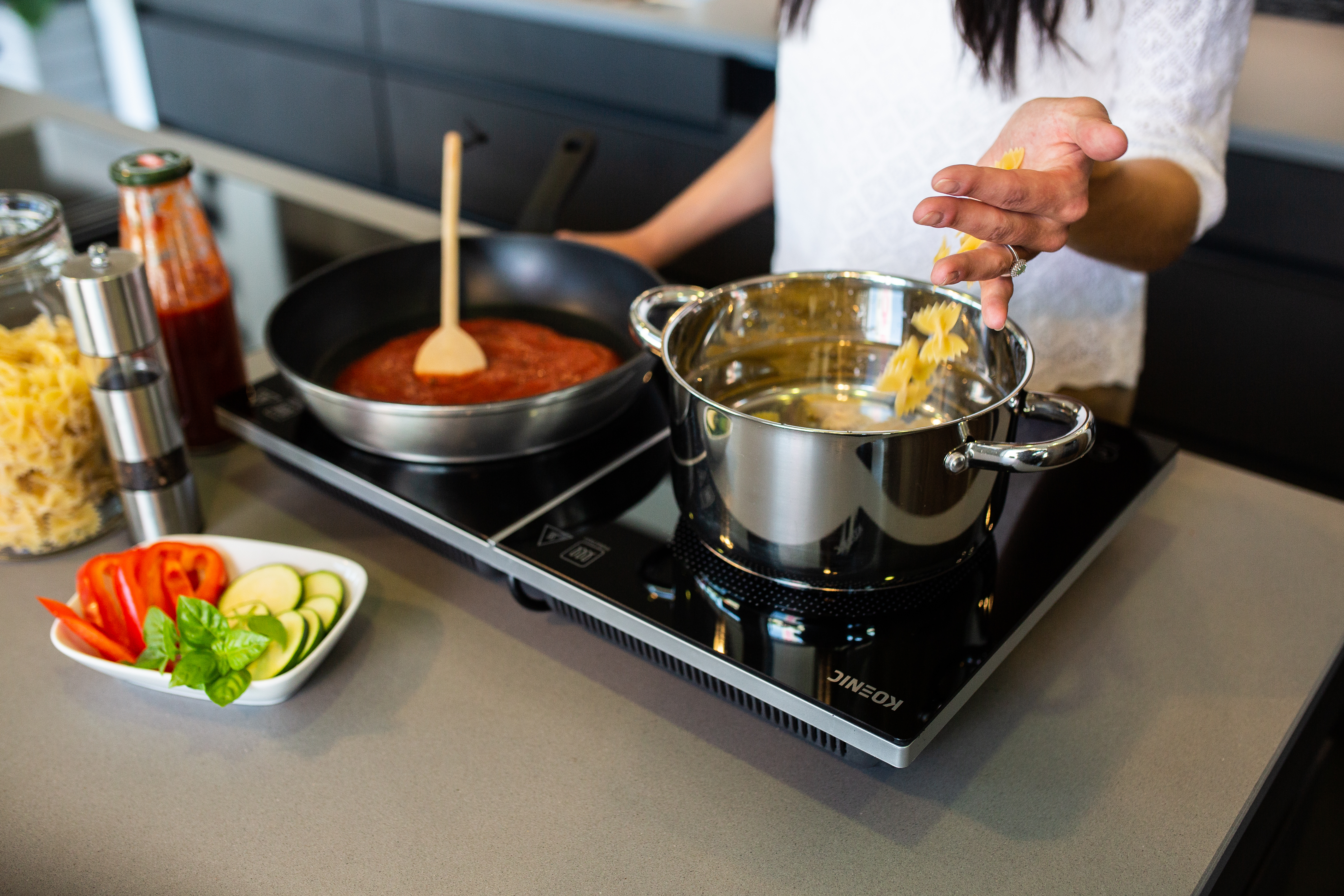 Pasta wird in einen Topf gegeben. Pfanne mit Soße, Gemüse und Zutaten liegen auf der Theke mit einer Person.
