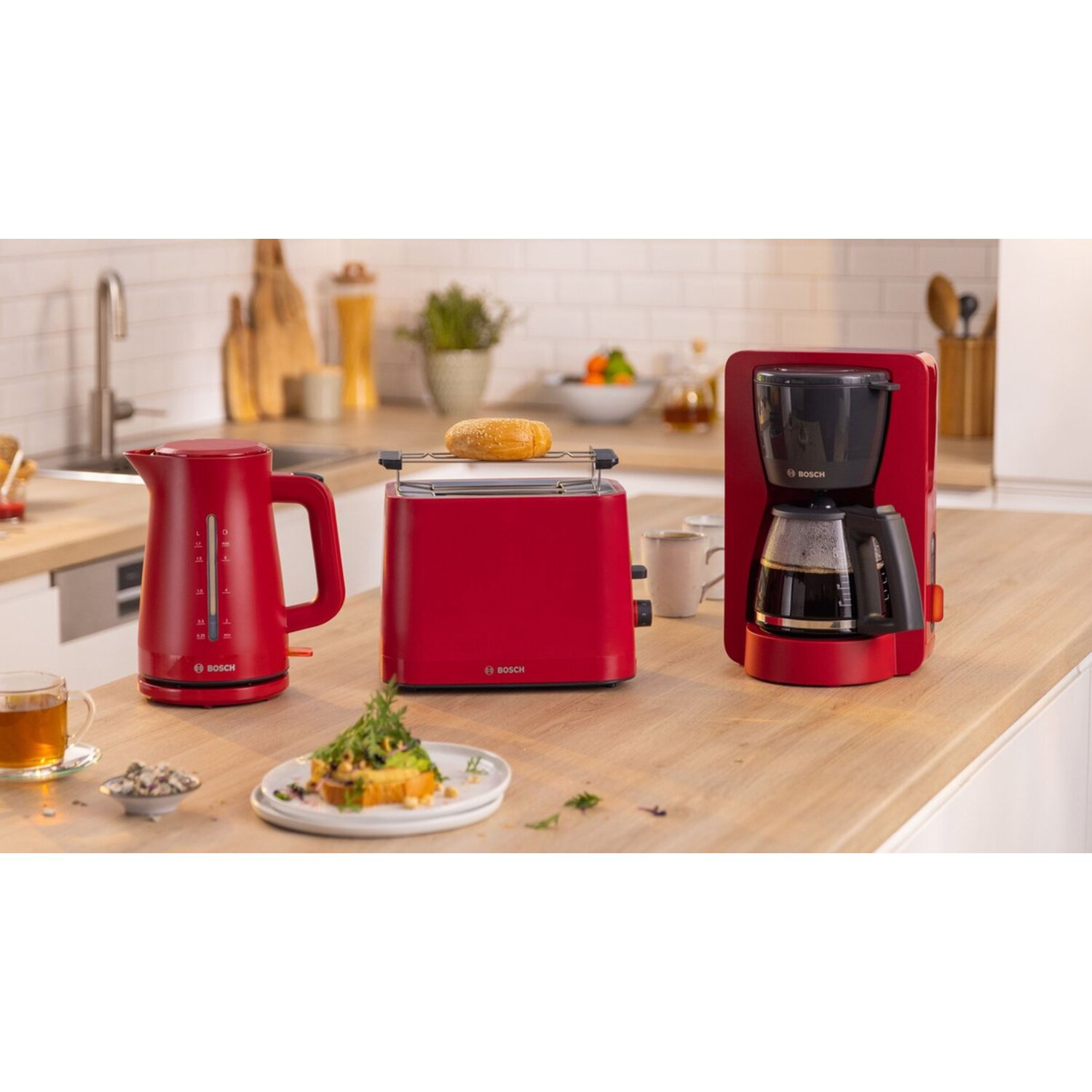 Red coffee maker, kettle, and toaster on a wooden surface. Bread with greens. Kitchen setting.