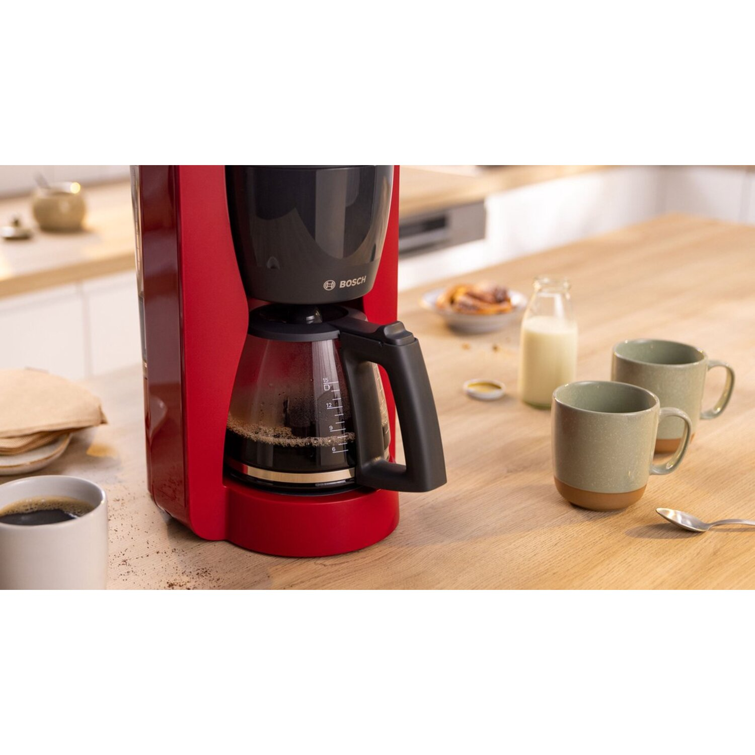 Red coffee machine, filled with coffee, with mugs and other items on a wooden counter.