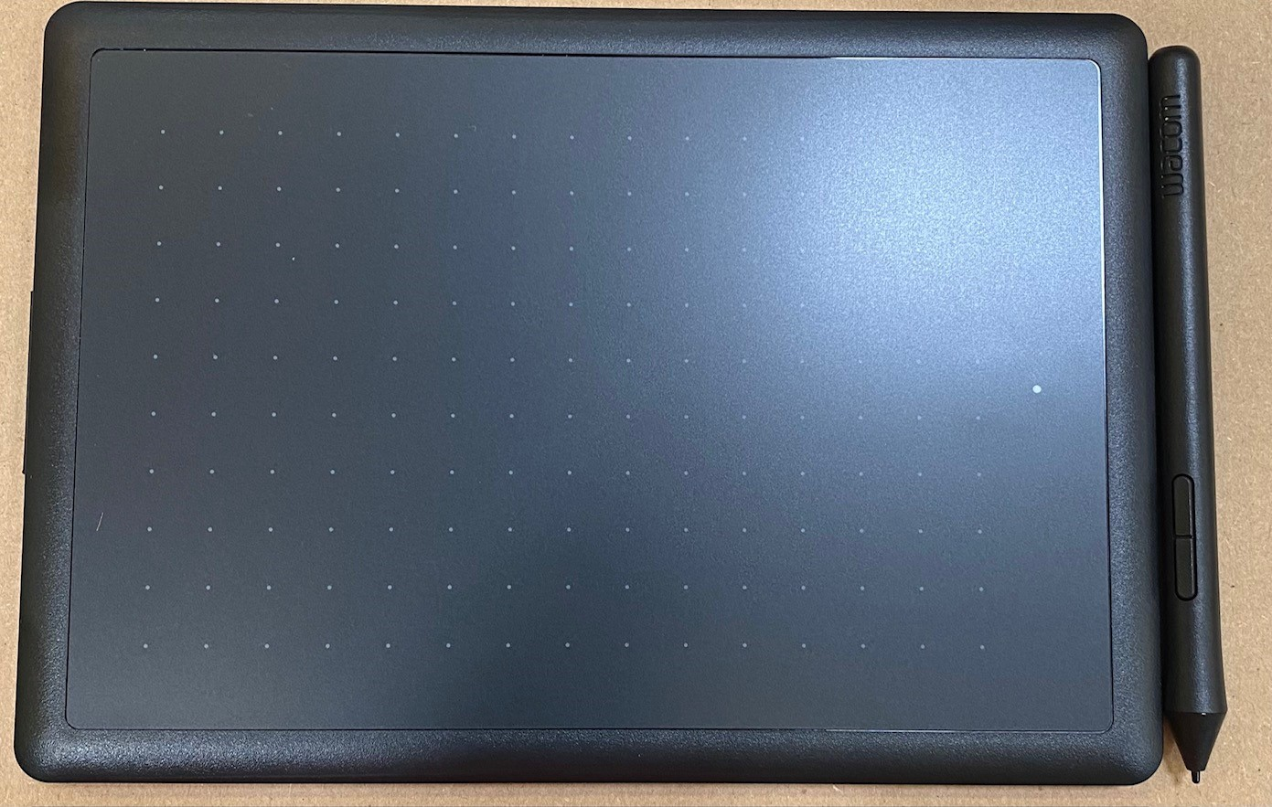 Czarny tablet graficzny Wacom z czarnym rysikiem. Tablet ma siatkę kropek i leży na brązowej powierzchni.