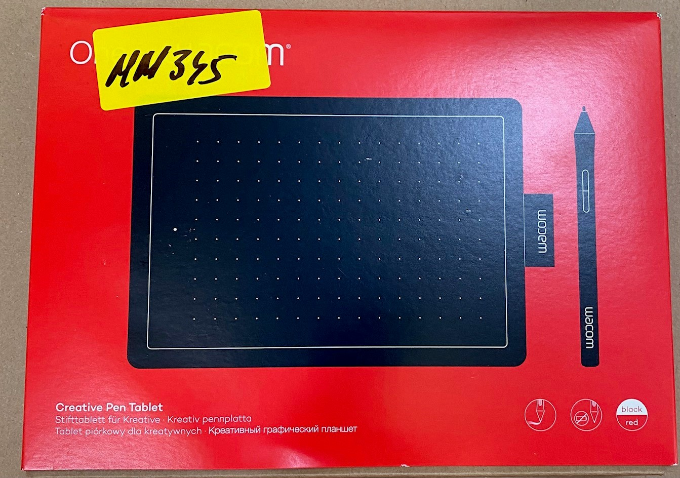 Czerwone pudełko zawiera czarny tablet i długopis. Żółta naklejka znajduje się na górze. Marka Wacom jest widoczna.