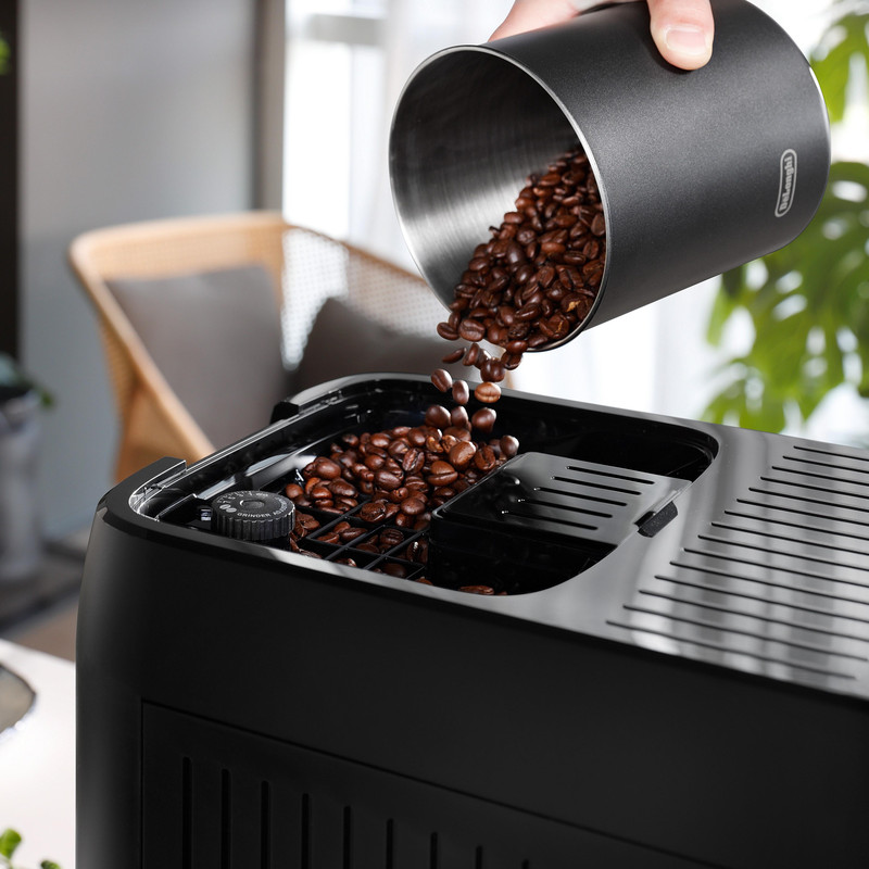 Ziarna kawy są wsypywane z czarnego pojemnika do czarnego ekspresu do kawy. W tle krzesło i roślina.