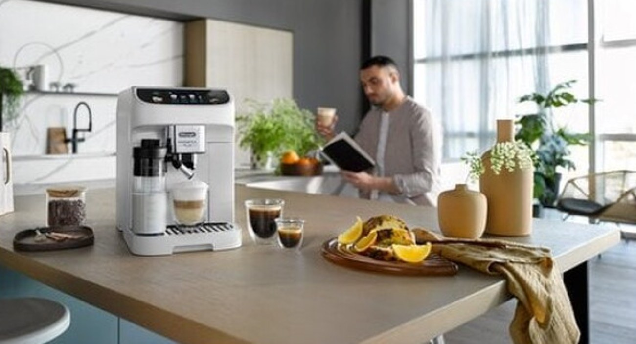 Ekspres do kawy, napoje i jedzenie na stole. Mężczyzna czyta książkę w tle. Obecne są również wazony i rośliny.