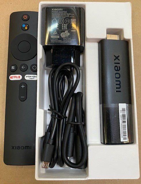 Czarny pilot Xiaomi, zasilacz, kabel i TV stick w białej tacy. Wszystkie elementy są czarne.