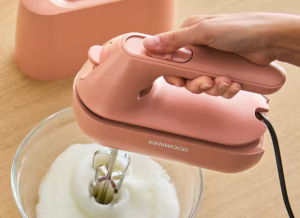 Ręka używa różowego miksera Kenwood do ubijania śmietany w szklanej misce. Różowy pojemnik do przechowywania jest w tle.