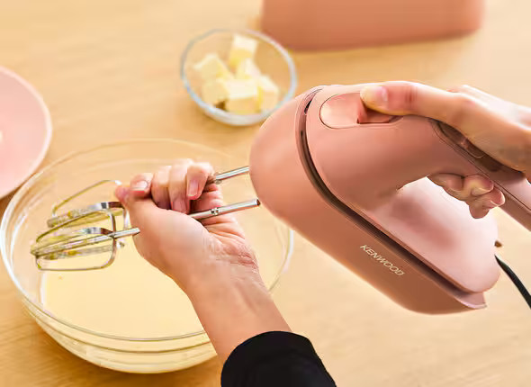 Osoba używa różowego miksera ręcznego do mieszania składników w szklanej misce. Widoczne są również masło i talerz.