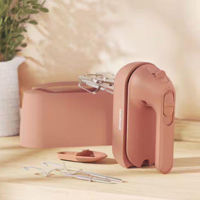 Różowy mikser ręczny Kenwood z nasadkami, etui i małym naczyniem na drewnianej powierzchni.