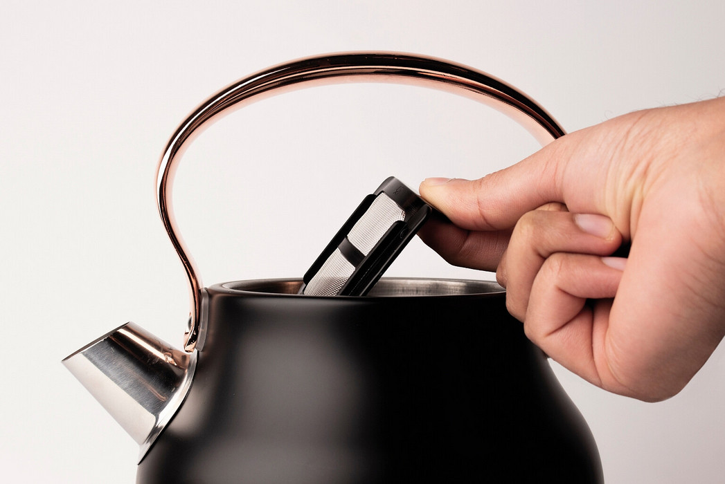 Ręka wkłada filtr do herbaty do czarnego czajnika z miedzianym uchwytem. Czajnik stoi na białym tle.