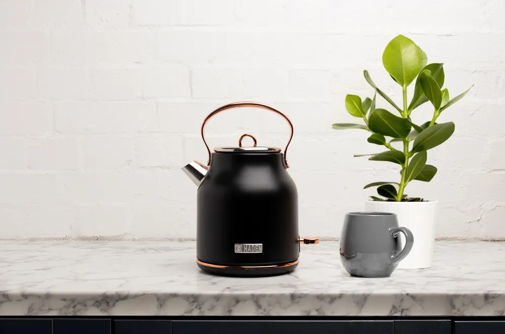 Czarny czajnik z miedzianymi akcentami, szary kubek i roślina doniczkowa na marmurowym blacie na tle białej ceglanej ściany.