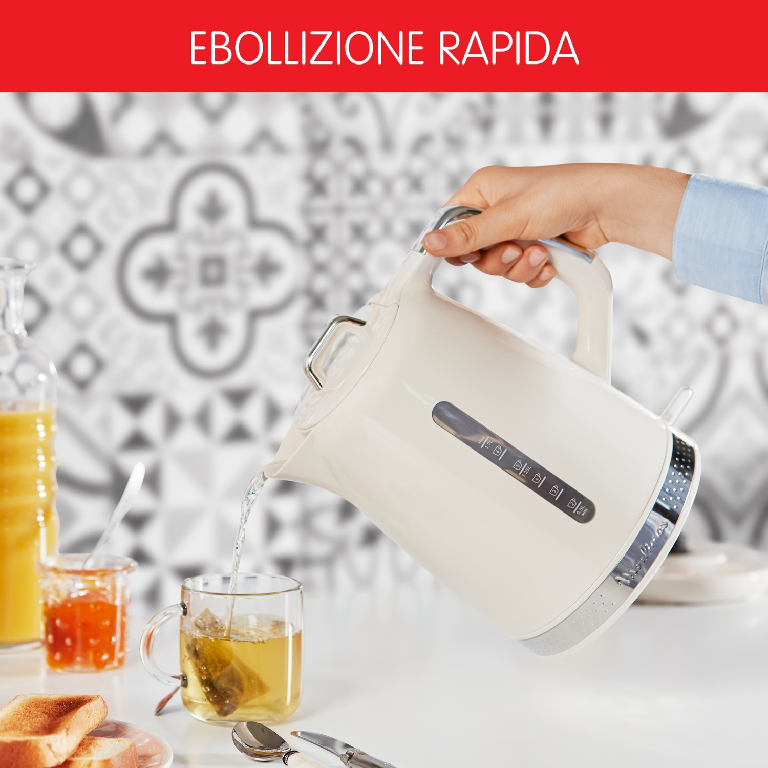 Una mano versa acqua da un bollitore bianco in un bicchiere di tè. Sono visibili anche toast, marmellata e una bottiglia.