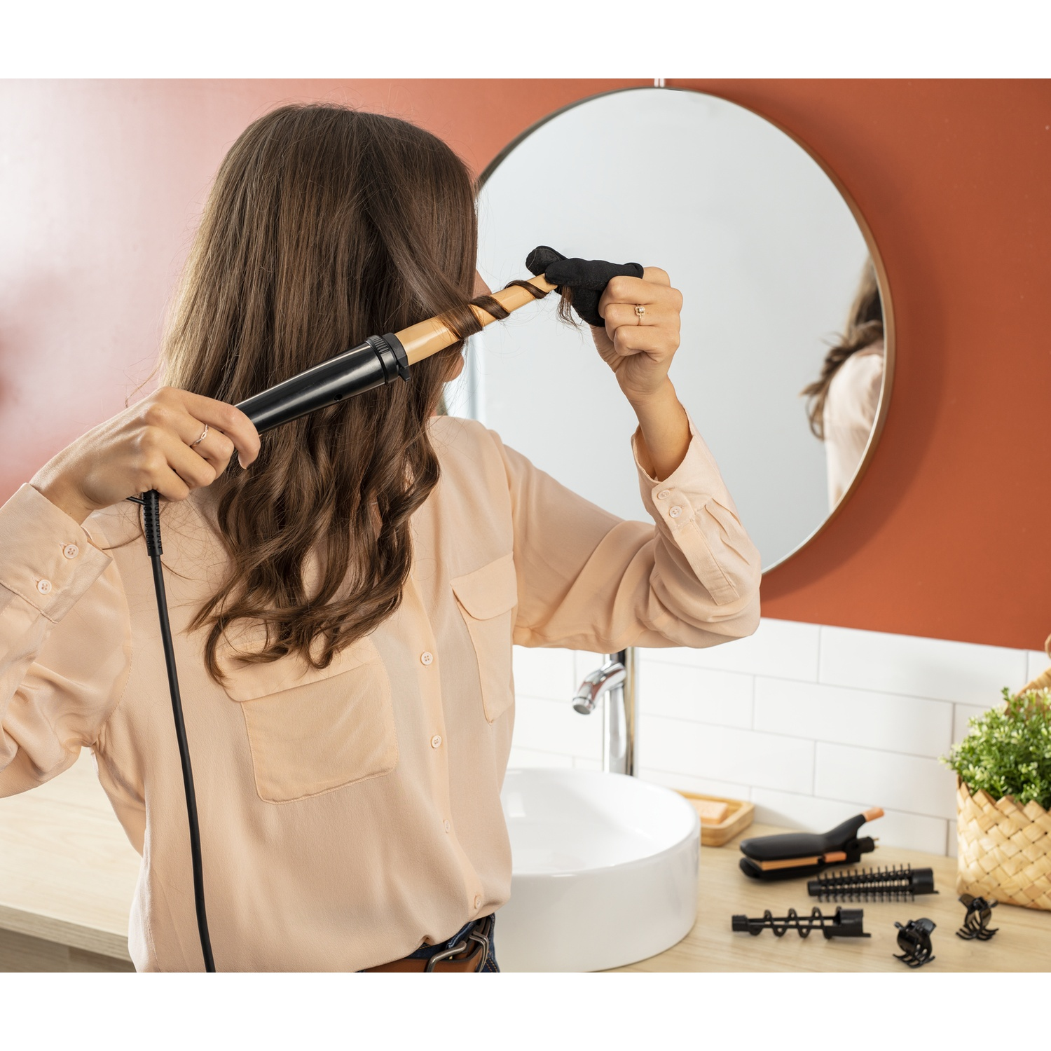 Donna che si arriccia i capelli con un ferro arricciacapelli davanti a uno specchio. Accessori sul bancone.