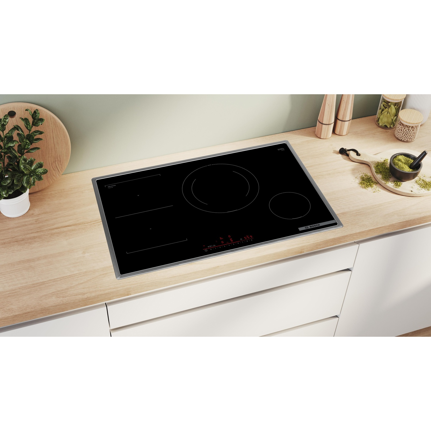 Piano cottura a induzione nero su un piano di lavoro in legno chiaro. Ambiente cucina con piante, barattoli e mortaio sullo sfondo.