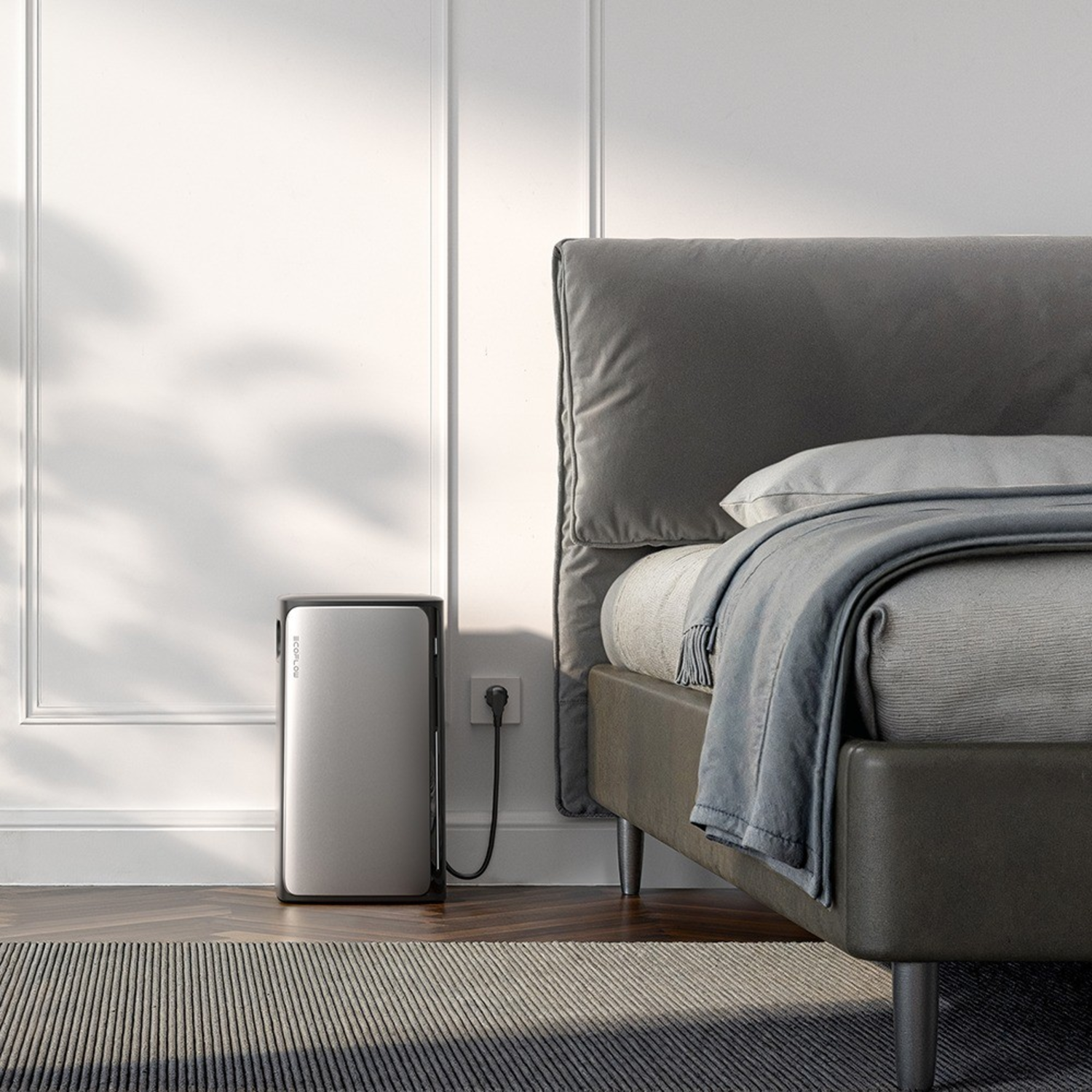 A battery storage system next to a bed. A rug is on the floor, and a wall is behind the bed.