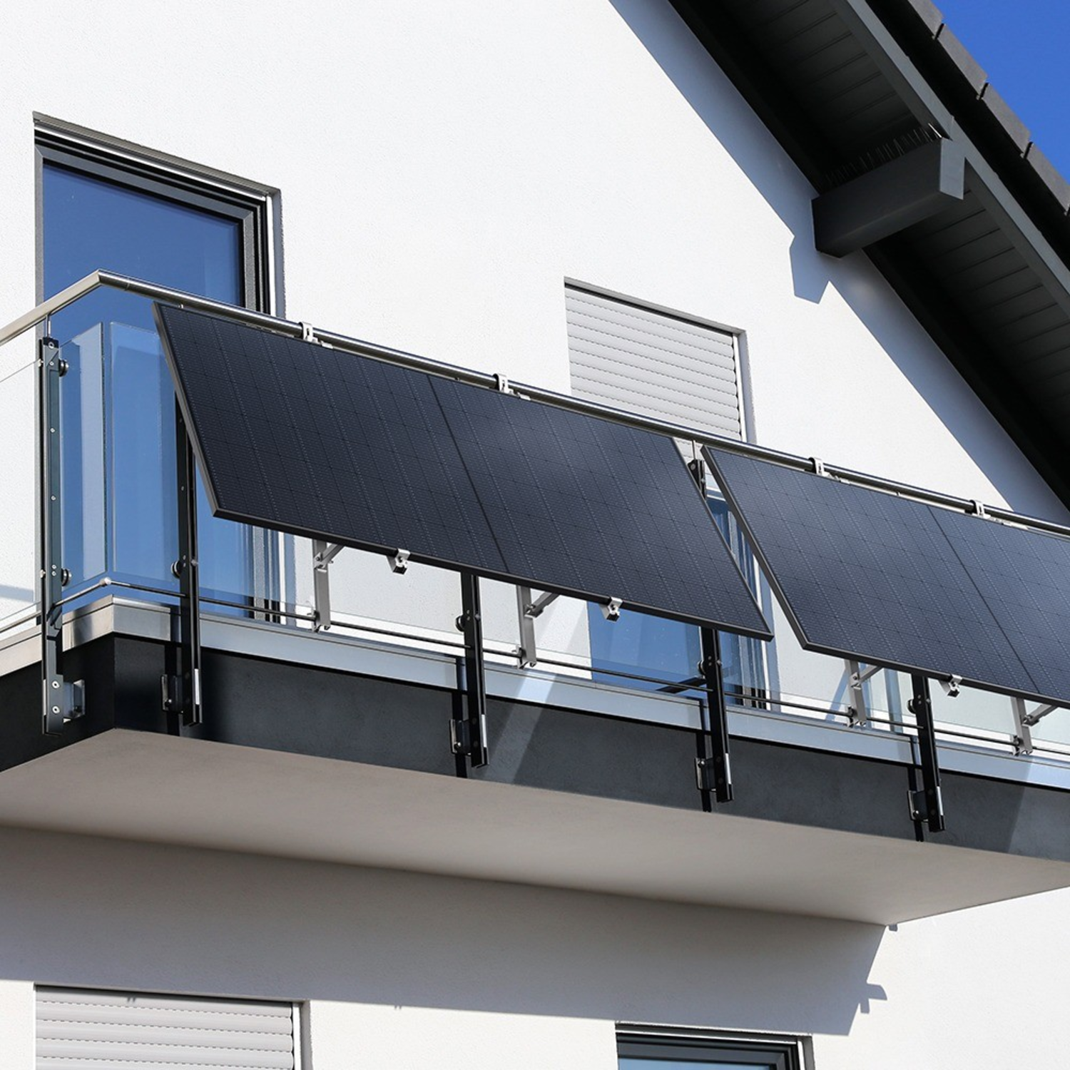 Panneaux solaires sur un balcon. Bâtiment blanc, balustrade en verre, cadres noirs. Ciel bleu visible.