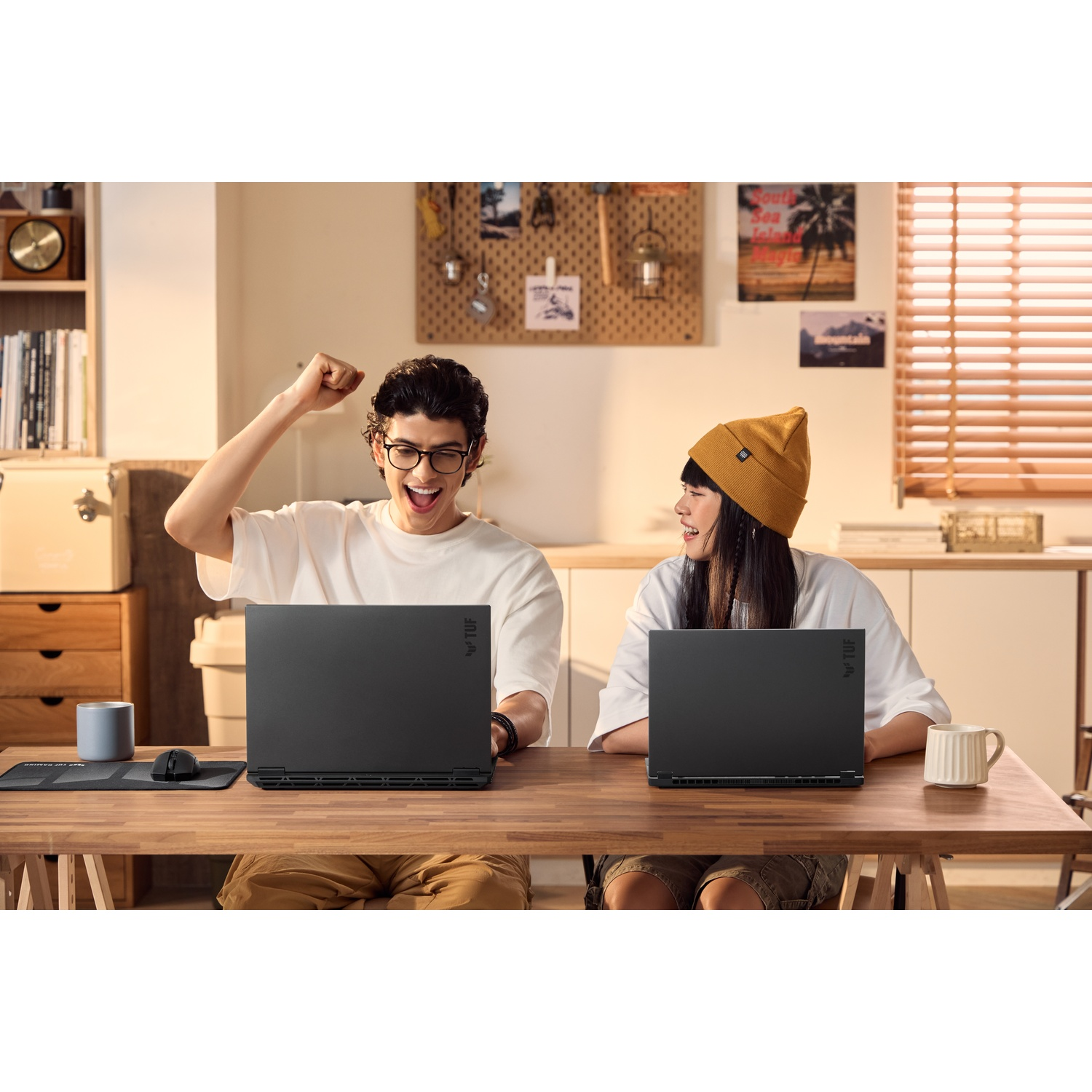 Due persone con laptop a un tavolo, una che esulta. Tavolo in legno, tazze, un mouse e una libreria visibili.