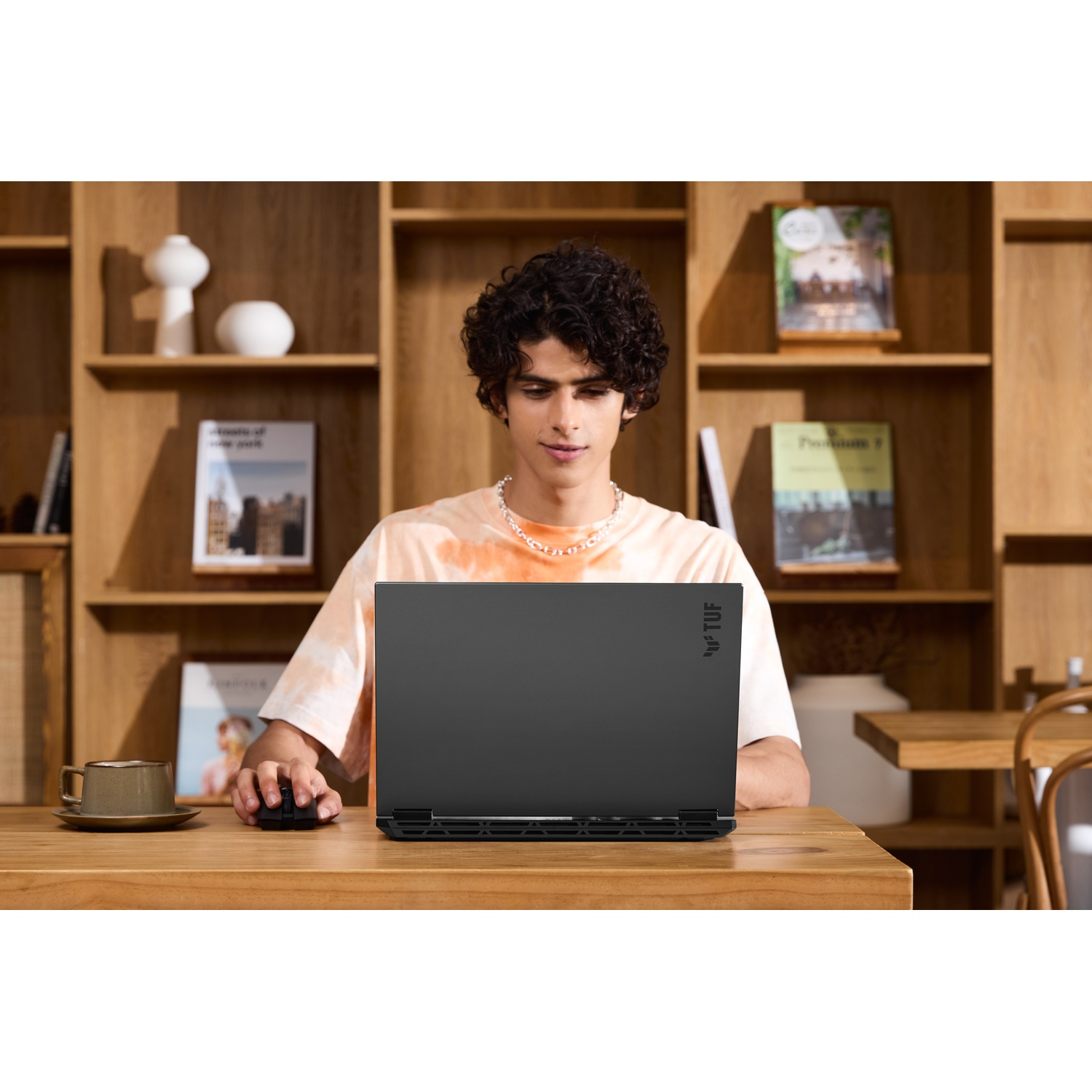 Uomo che usa un laptop. Tavolo di legno, tazza, mouse e libreria sullo sfondo.