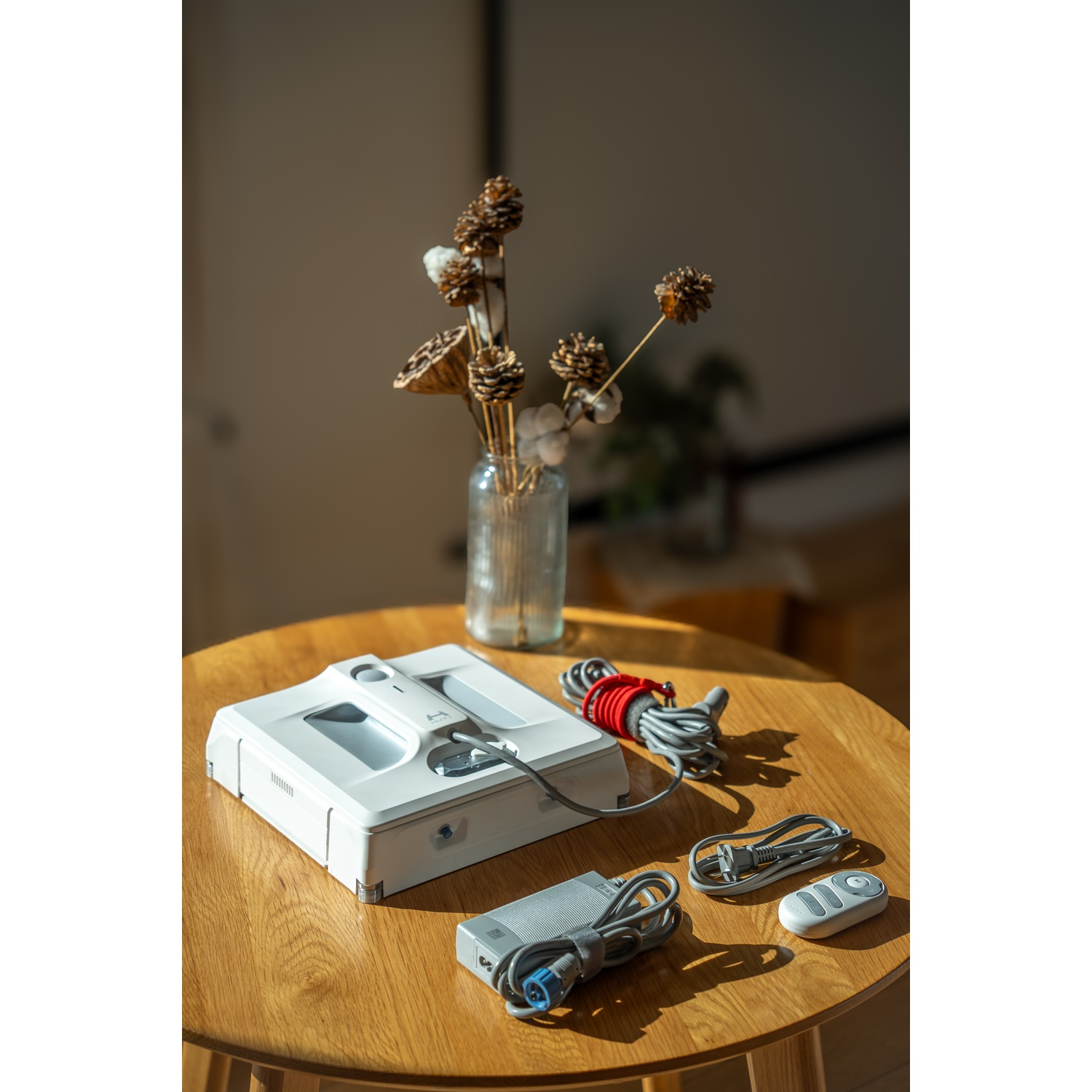 Robot per la pulizia dei vetri bianco su tavolo di legno, con accessori e vaso di fiori secchi. Luce solare, sfondo sfocato.