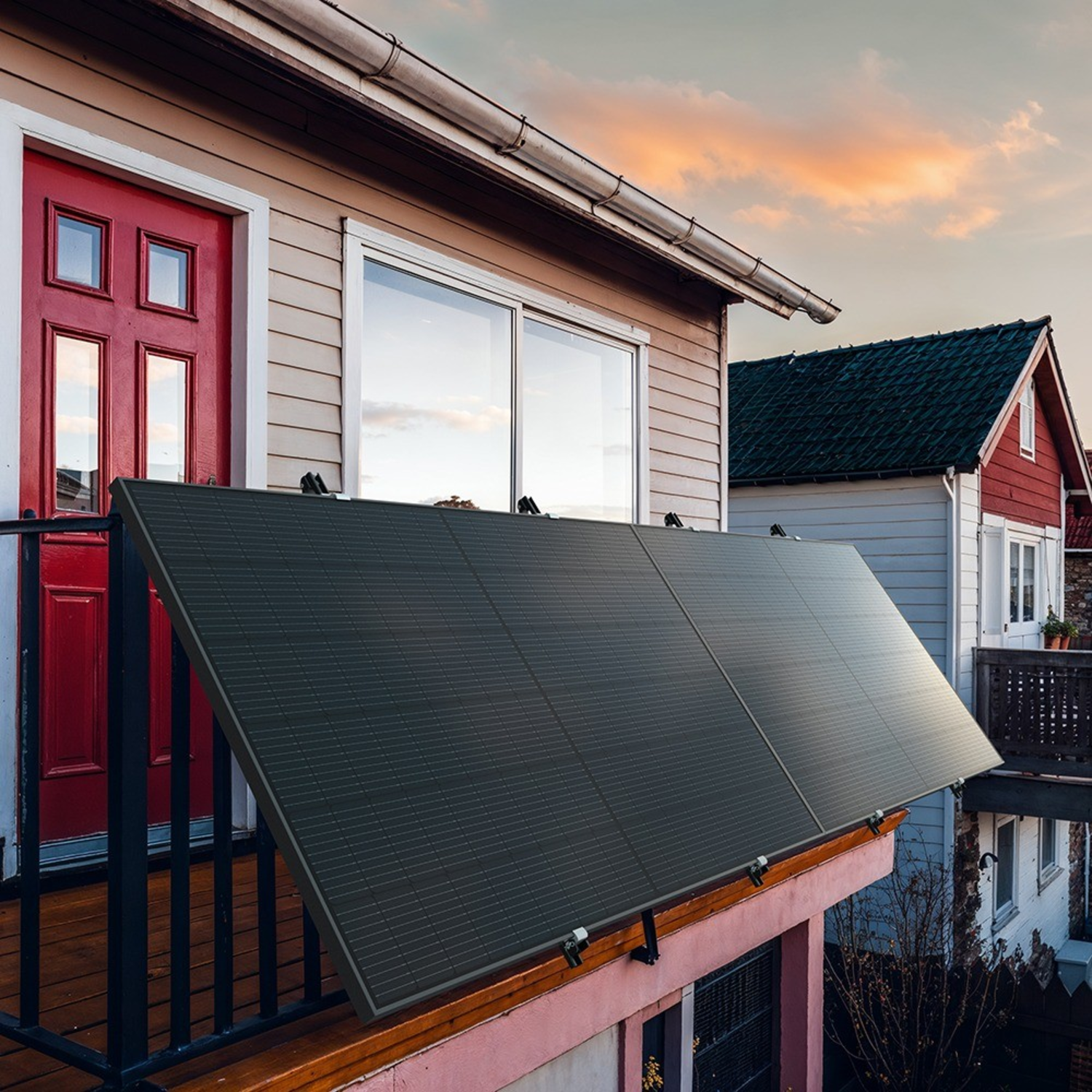 Solar panels on a balcony of a house, red door, other houses in the background.