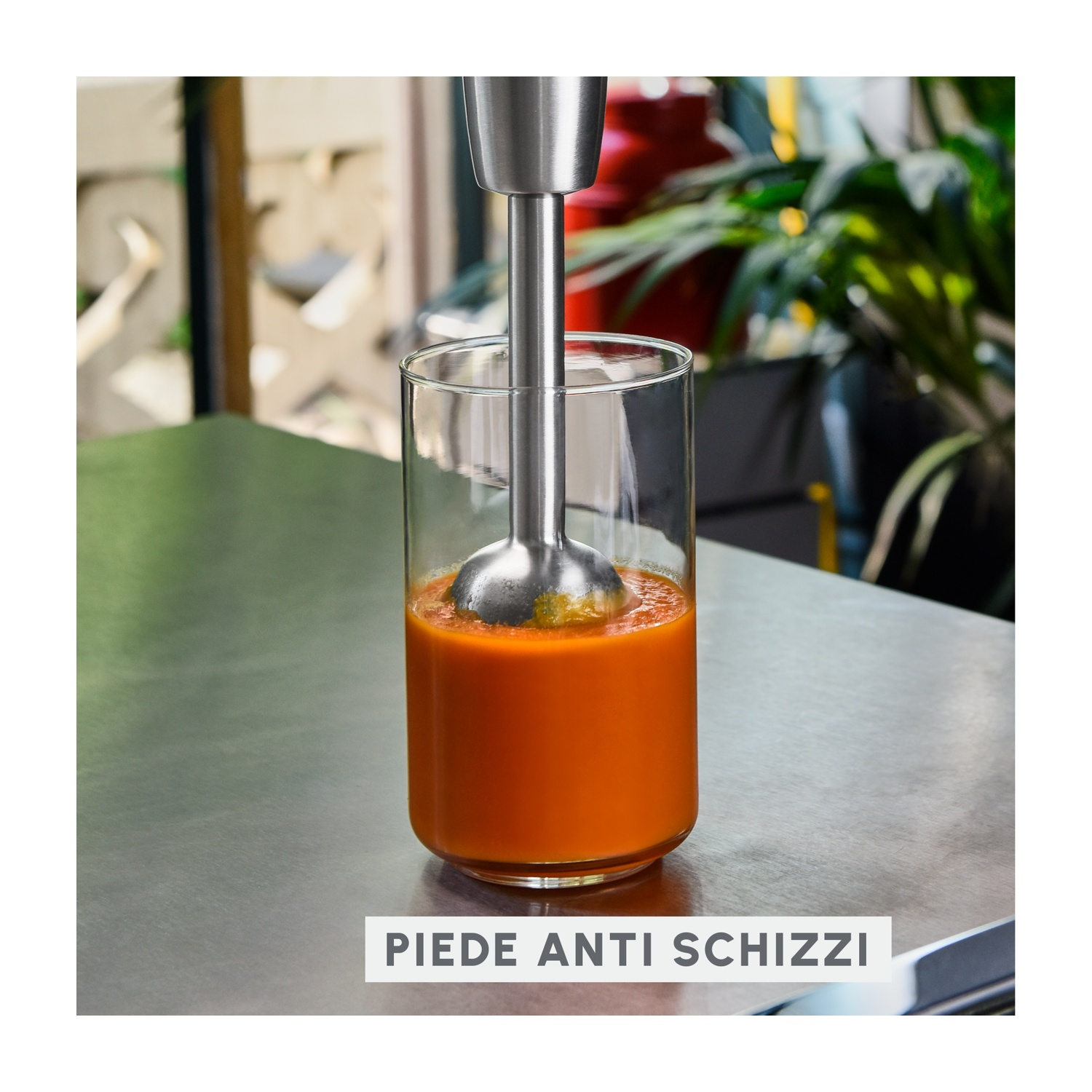 Un frullatore a immersione in un bicchiere di liquido arancione. Il frullatore è in acciaio inossidabile. Lo sfondo ha delle piante.