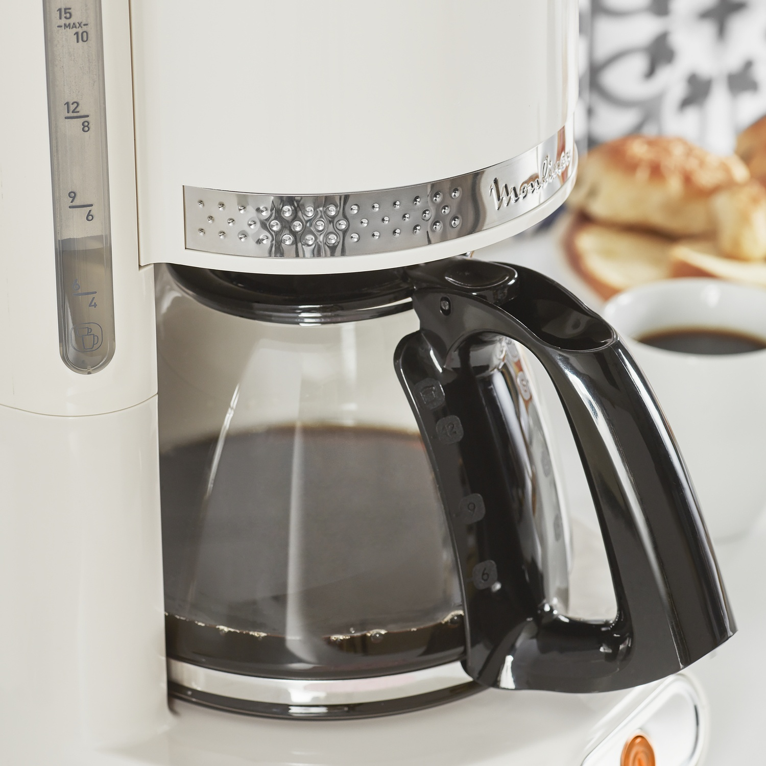 Primo piano di una caffettiera con una caraffa di vetro piena di caffè, una tazza e pane su una superficie bianca.