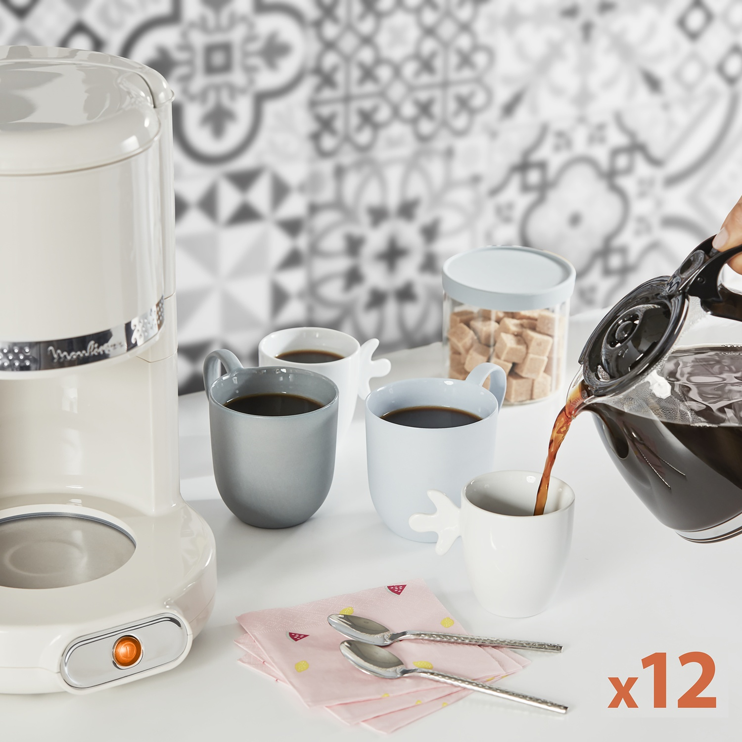 Il caffè viene versato da una caraffa in una tazza bianca. Altre tazze, zucchero e una macchina per il caffè sono su un tavolo.