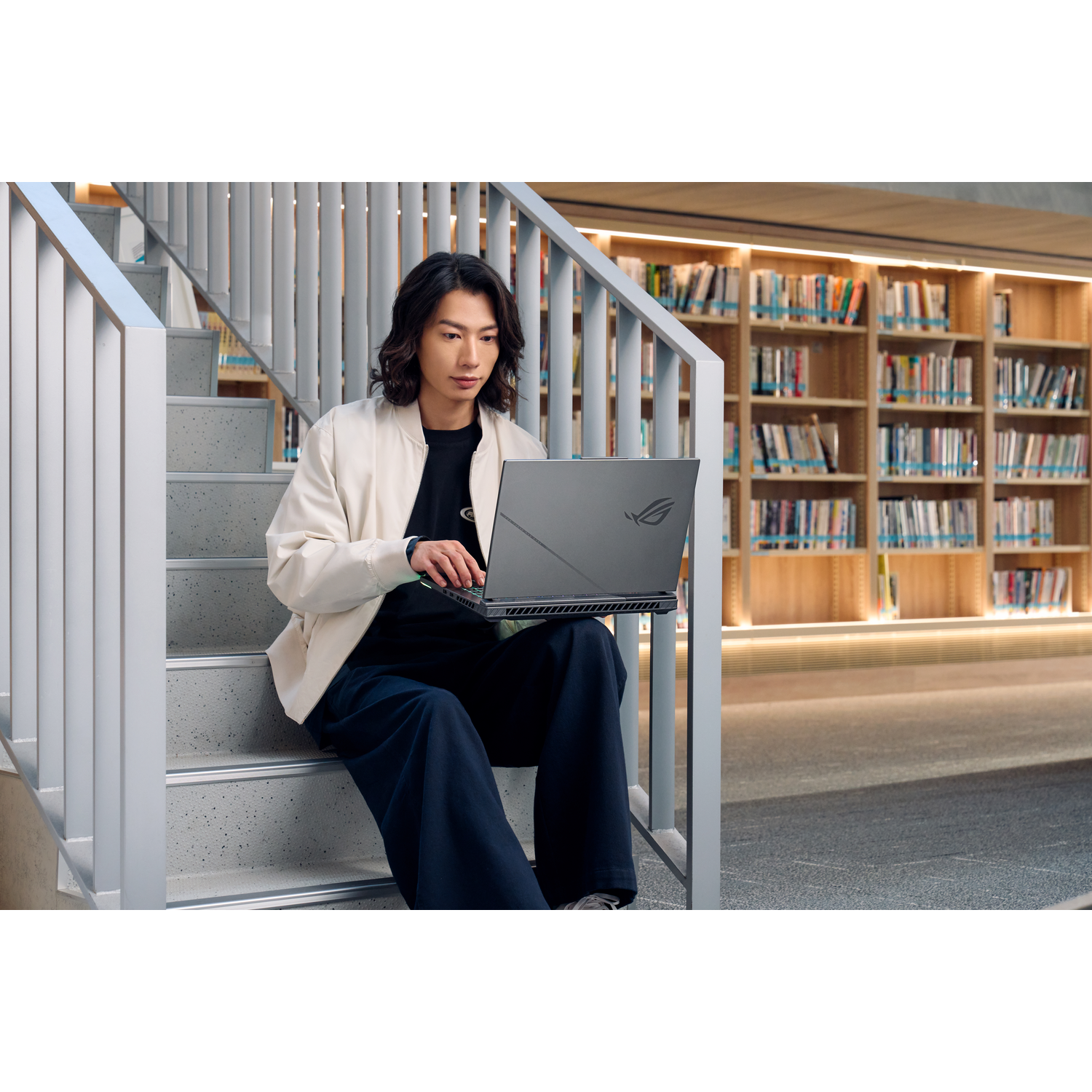 Persoon die een laptop gebruikt op een trap in een bibliotheek. Boekenrekken op de achtergrond.