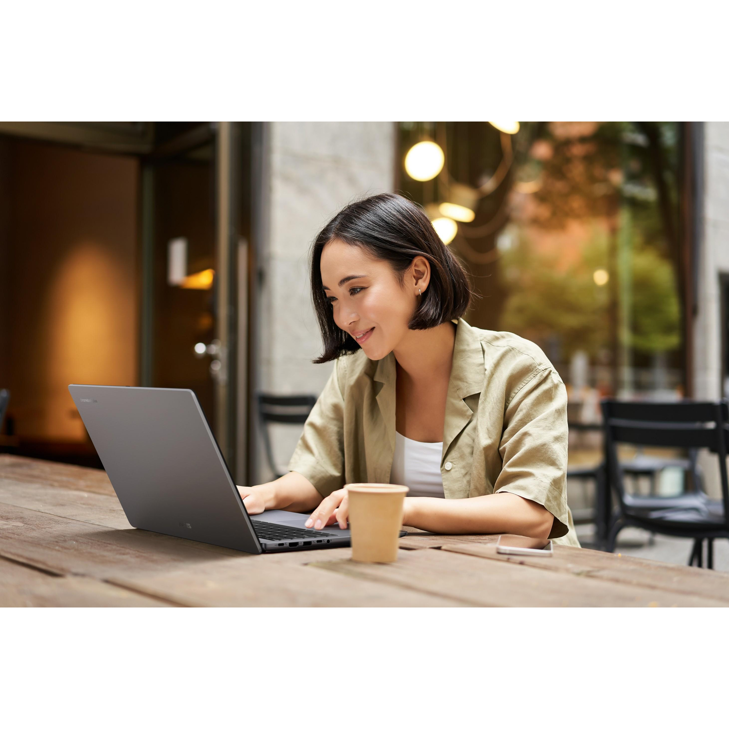 Woman using a dark gray laptop at a wooden table, with a coffee cup and phone.