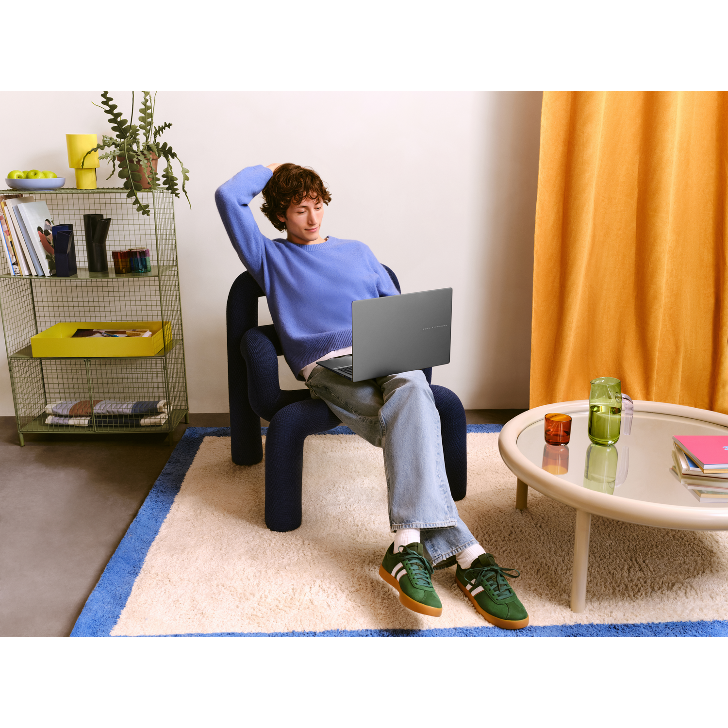 Man sitting in a chair using a laptop, wearing a blue sweater. Colorful background with decor.