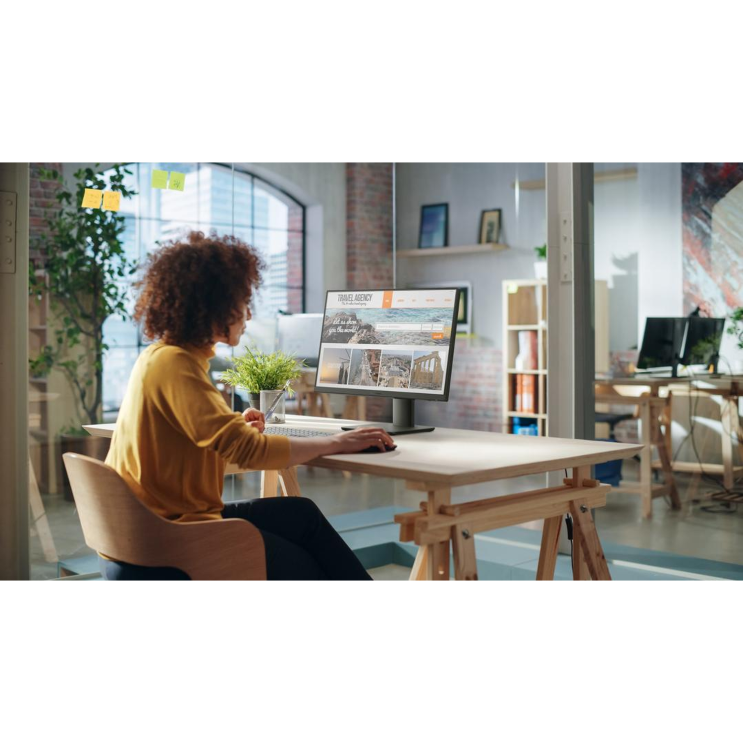 Een vrouw werkt aan een bureau met een computermonitor die een website weergeeft, in de buurt van een raam en planten.