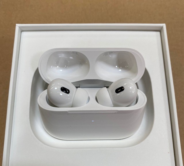 Białe AirPods Pro Apple w otwartym etui. Tło: białe pudełko na brązowej powierzchni.