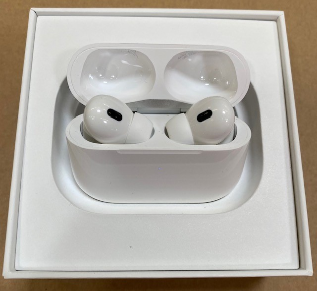 Białe AirPods Pro w otwartym etui ładującym, wewnątrz białego pudełka, na brązowej powierzchni.