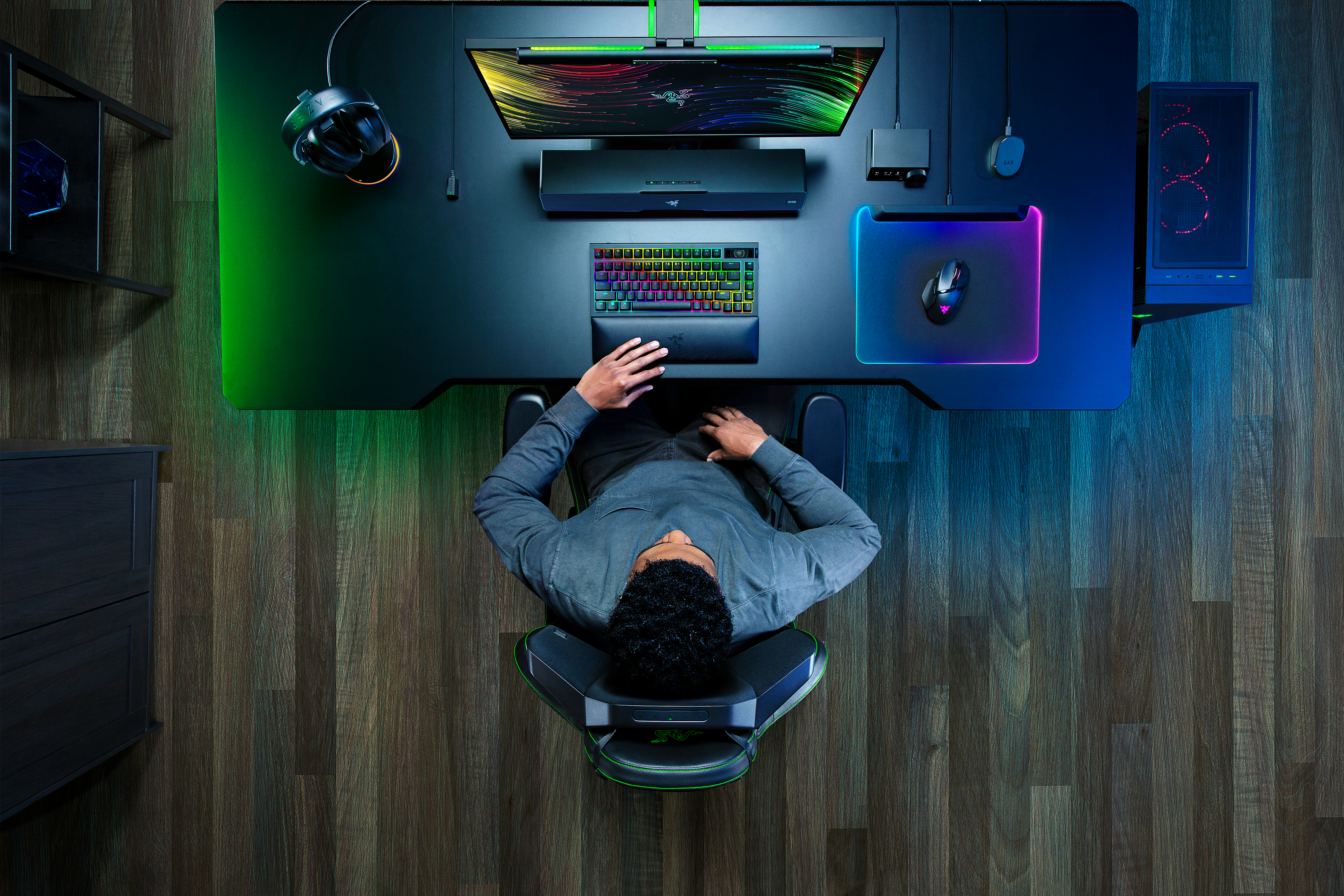 Draufsicht einer Person, die an einem Gaming-Schreibtisch mit Monitor, Tastatur, Maus und Headset sitzt. Der Raum hat dunklen Holzboden.