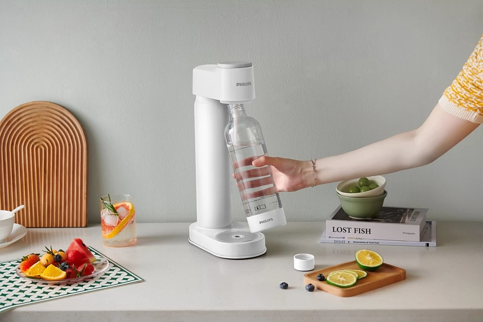 Kobieta wlewa wodę z urządzenia do produkcji sody do szklanki, na stole owoce.