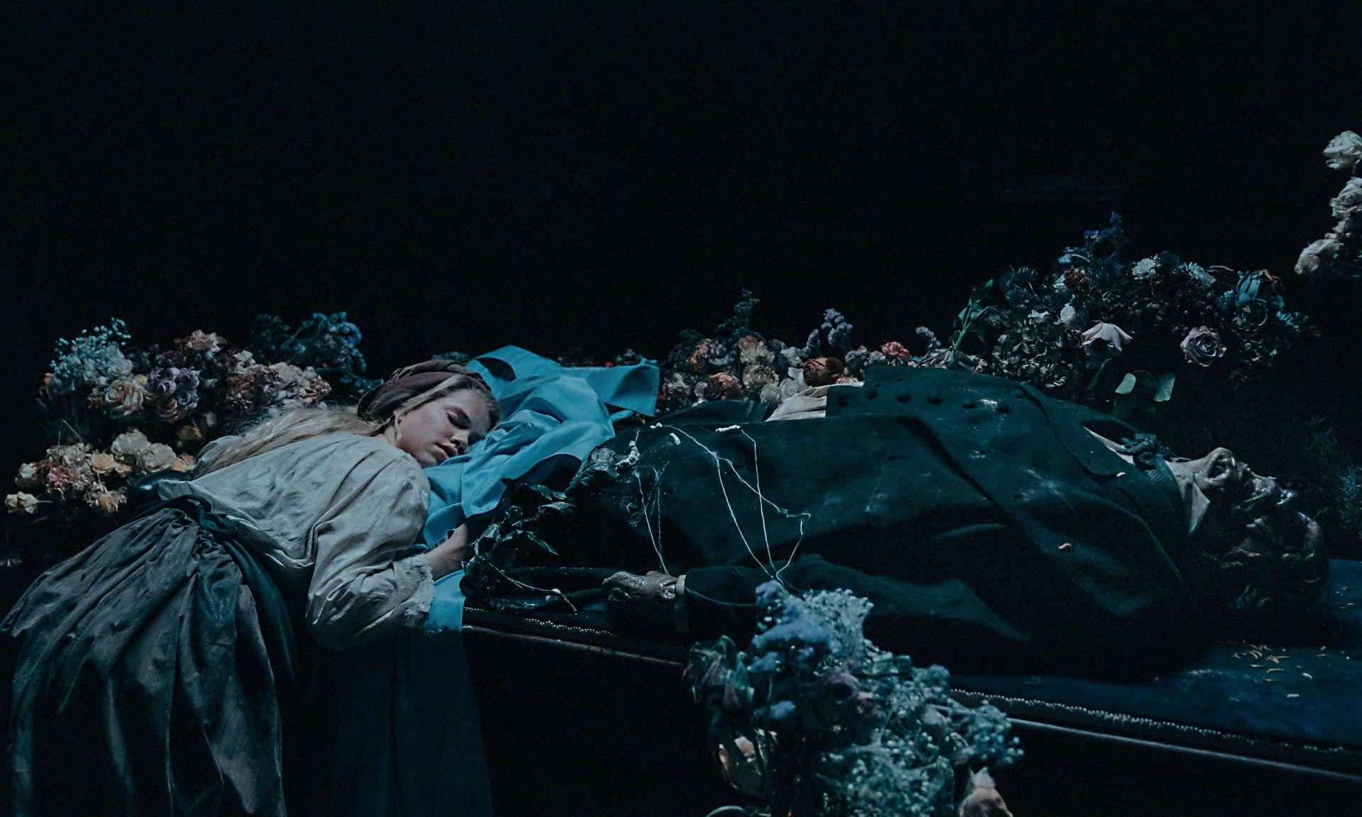 Eine Frau trauert über einem Mann, der auf einem Tisch liegt, umgeben von Blumen, in dunkler Umgebung.