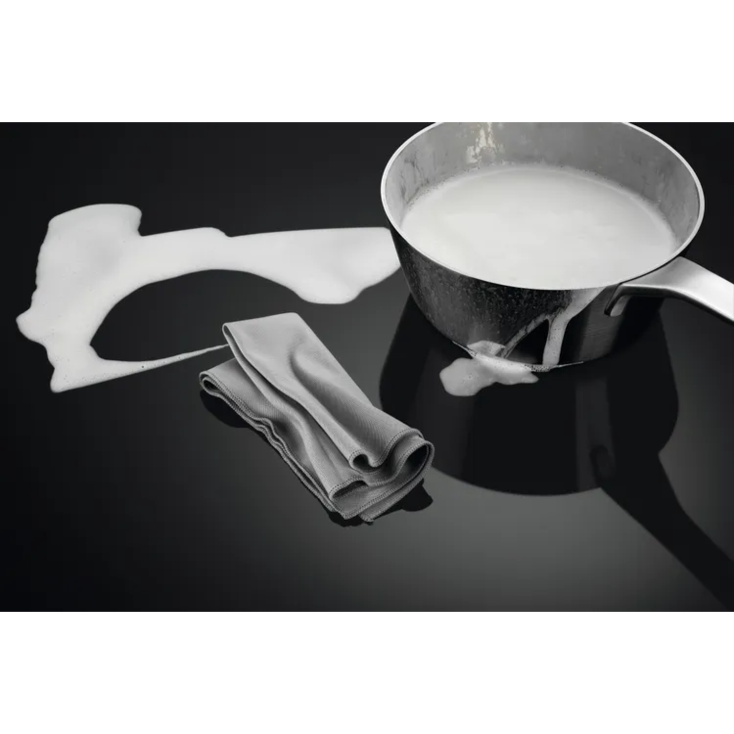 Pot with spilled milk on a black cooktop. A gray cloth is next to the spill.