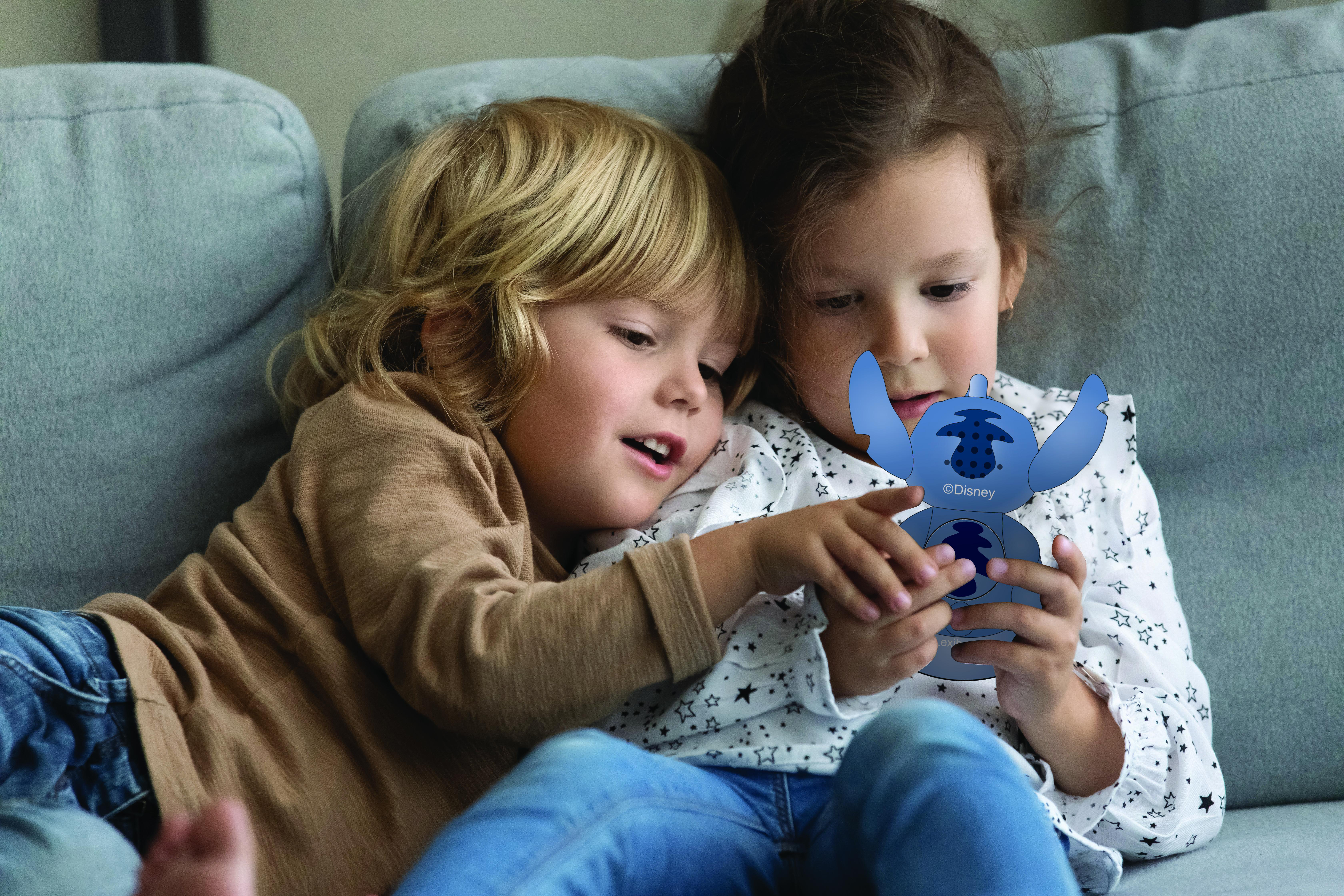 Zwei Kinder betrachten gemeinsam ein Stitch-Spielzeug auf einem grauen Sofa.