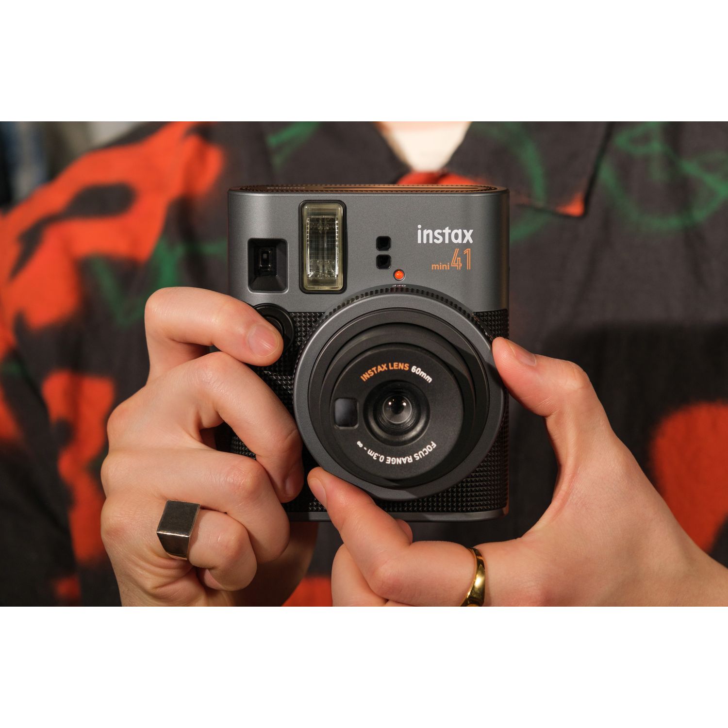 A person holds a gray Instax Mini 41 camera. The background is a person in a patterned shirt.