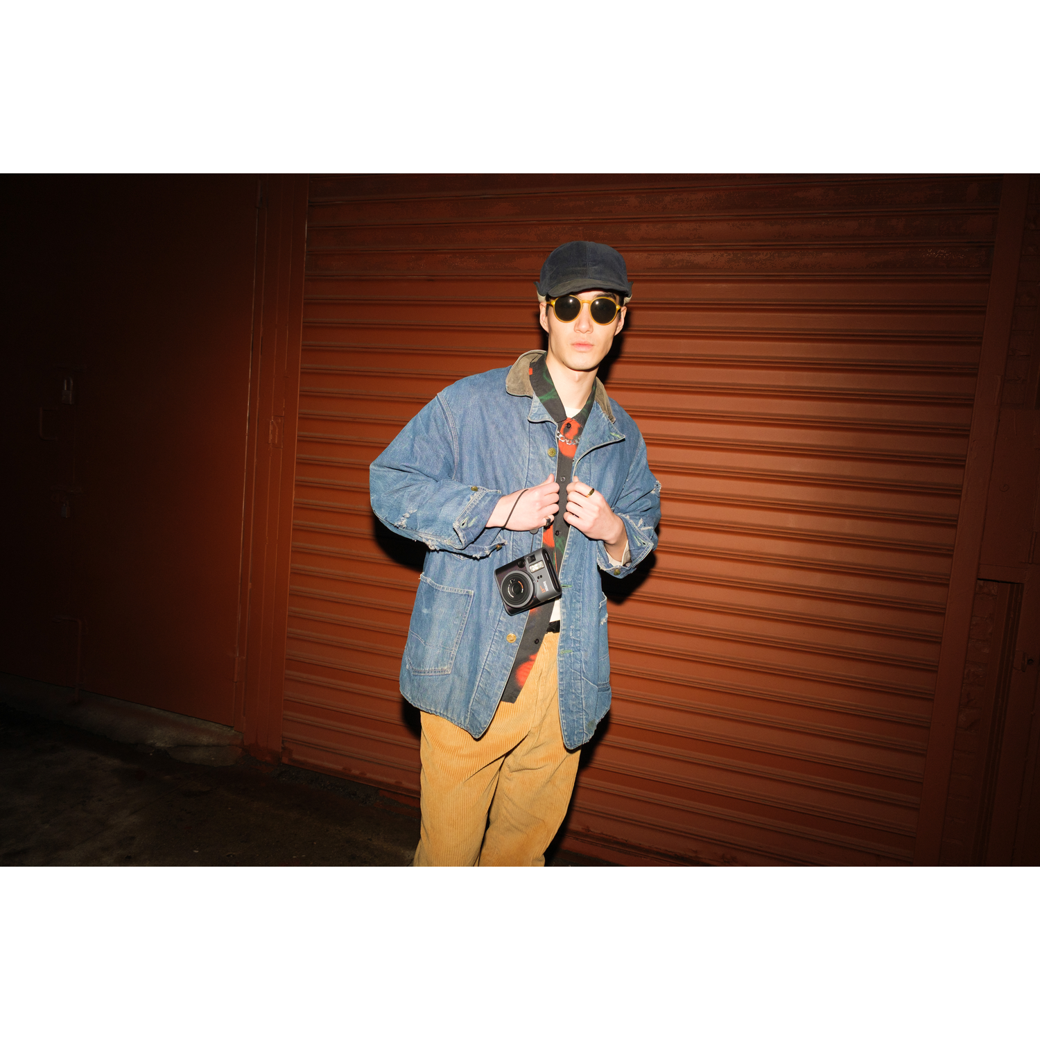 A person in a denim jacket and hat stands in front of a red shutter door holding a camera.