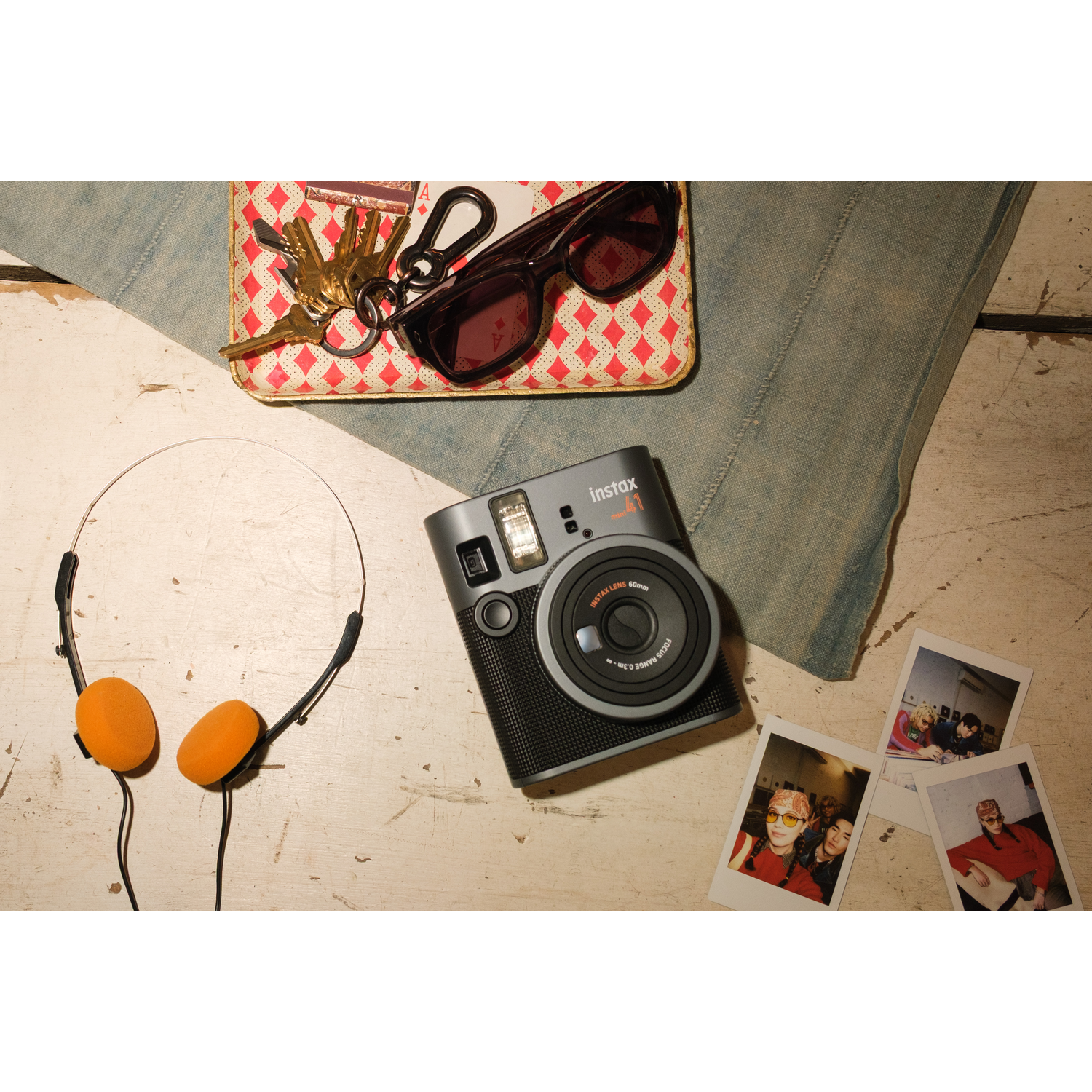 An Instax Mini 41 camera sits on a white table with keys, sunglasses, and headphones.