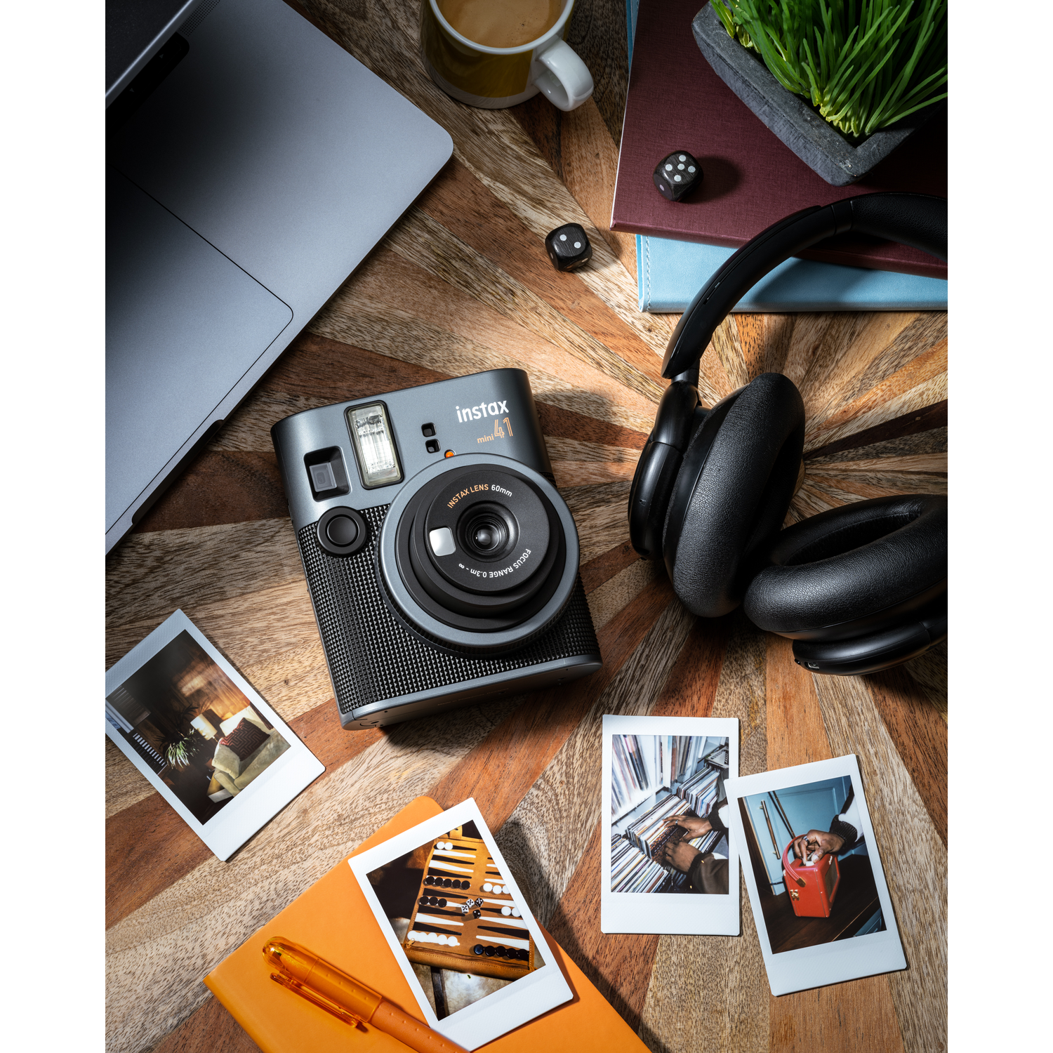 An Instax Mini 41 camera sits on a wooden table with headphones, a laptop, and photos.