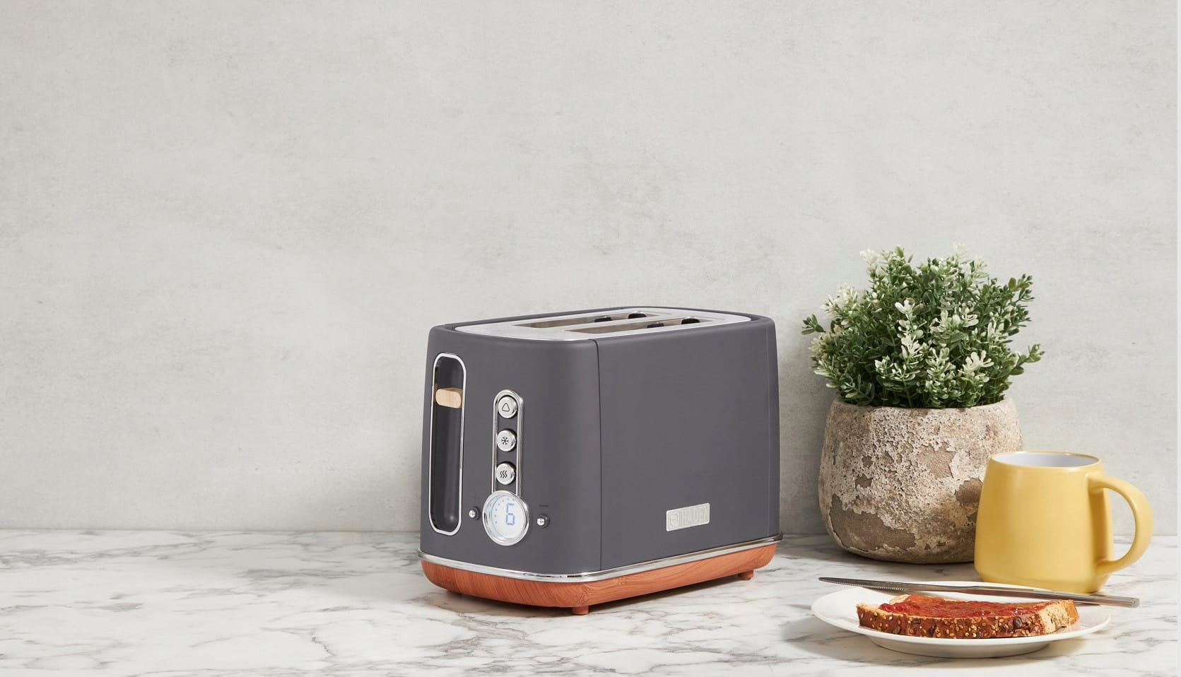 Szary toster z drewnianymi akcentami na marmurowym blacie, obok rośliny, żółtego kubka i tostu z dżemem.