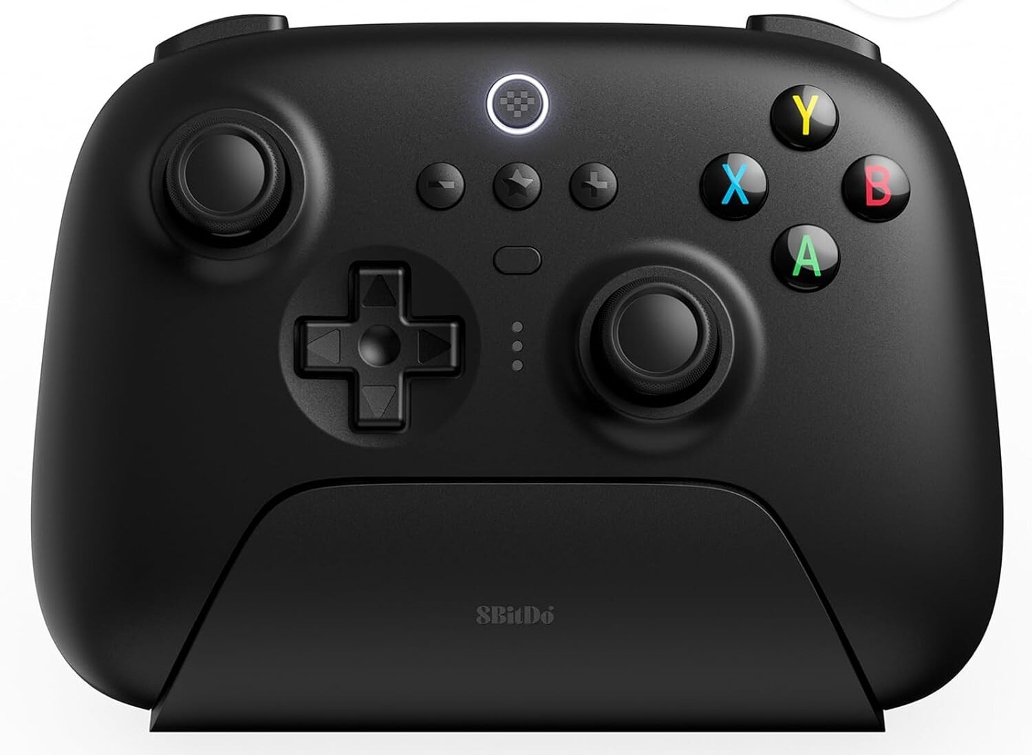 Czarny gamepad z kolorowymi przyciskami. Ma krzyżak i dwa joysticki. Pad jest na czarnej podstawce.