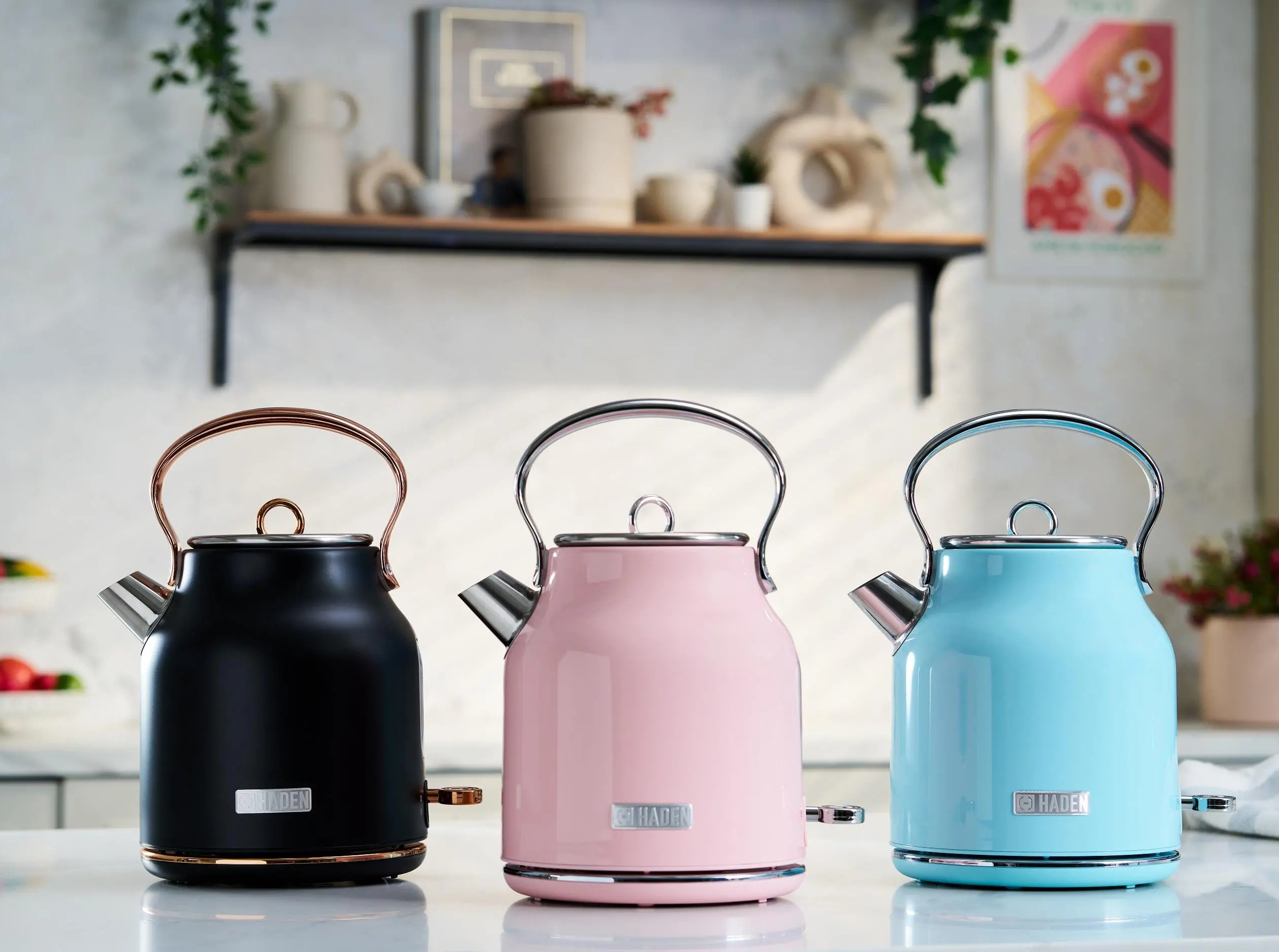 Trzy czajniki, czarny, różowy i niebieski, na powierzchni, z półką kuchenną w tle.