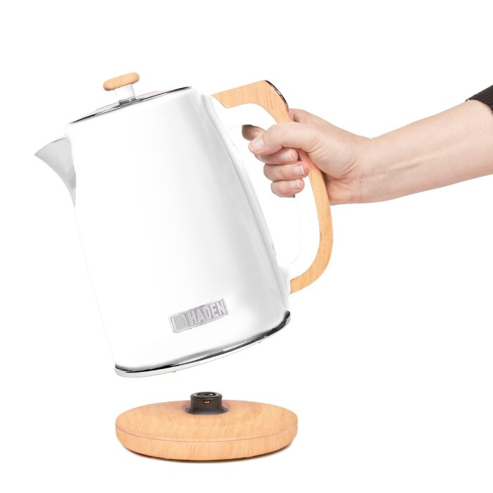 Biały czajnik elektryczny z drewnianym uchwytem trzymany jest nad drewnianą podstawą przez rękę. Na czajniku znajduje się napis 'HADEN'.