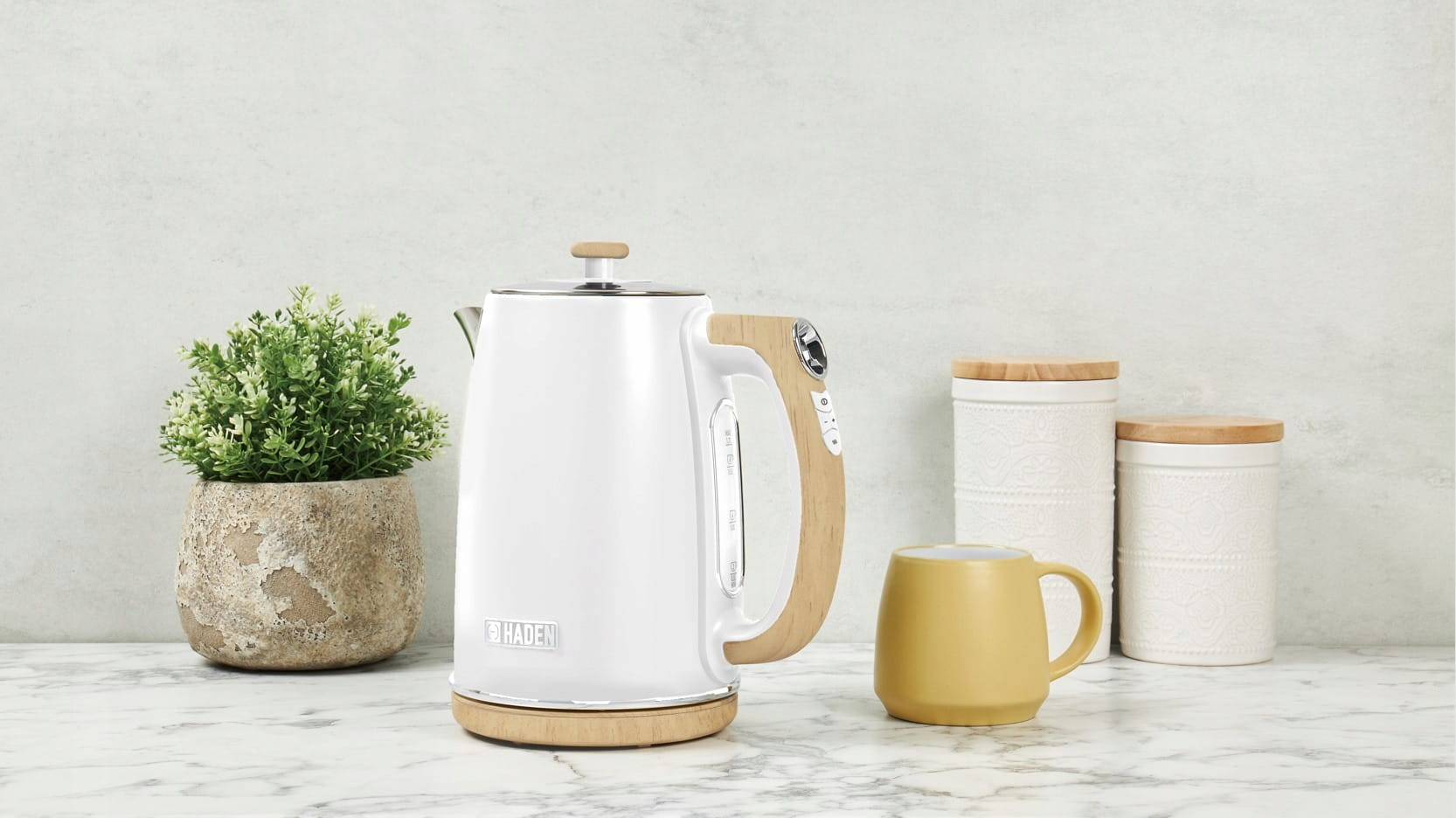 Biały czajnik elektryczny z drewnianymi akcentami, żółty kubek, roślina i ozdobne słoiki na marmurze.