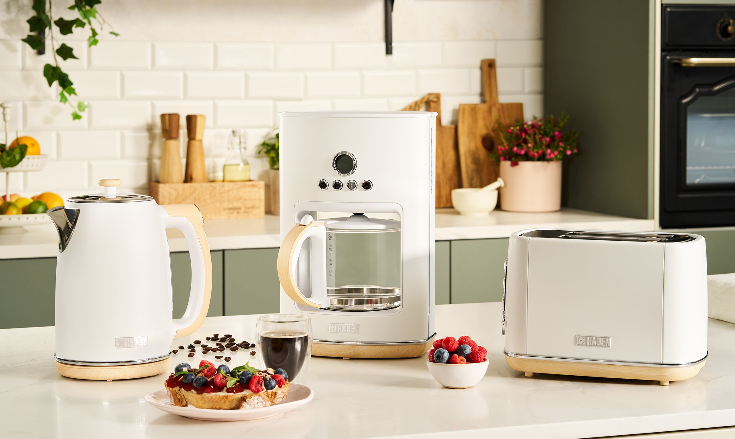 Scena kuchenna z białymi urządzeniami, tostami i kawą. Zawiera czajnik, ekspres do kawy, toster i przekąskę z jagodami.