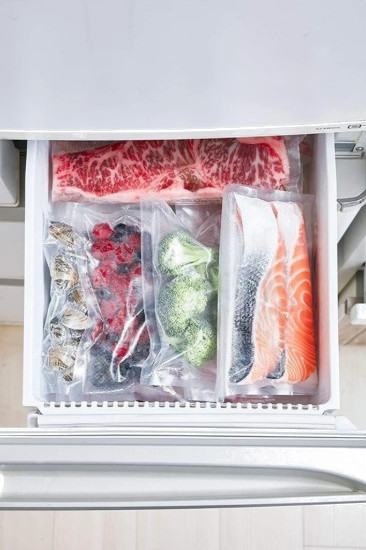 Otwarta szuflada lodówki z różnymi produktami pakowanymi próżniowo. Zawiera mięso, jagody, brokuły i ryby.