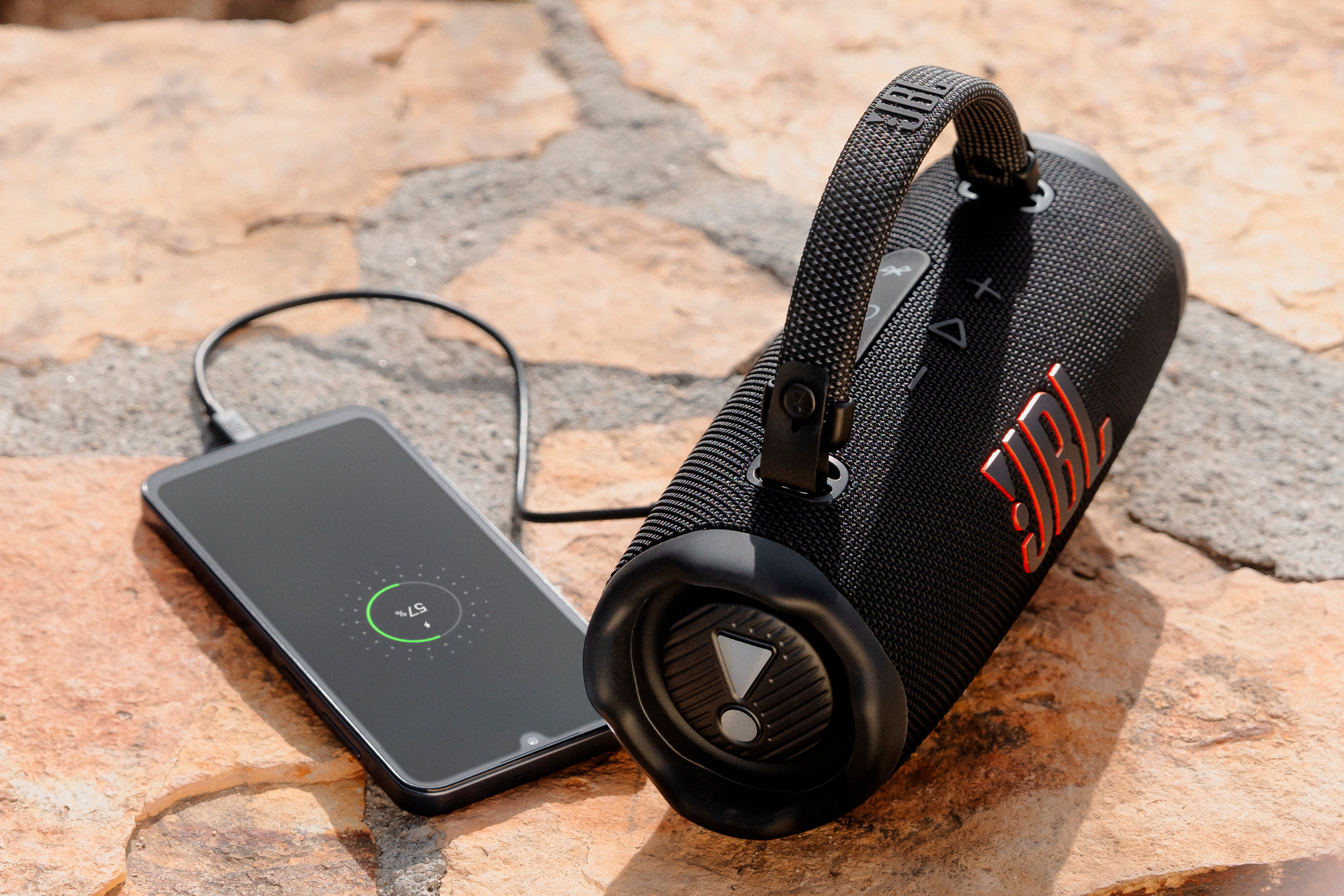Schwarzer JBL-Lautsprecher lädt ein Telefon. Das Telefon ist schwarz und mit einem Ladekabel mit dem Lautsprecher verbunden.