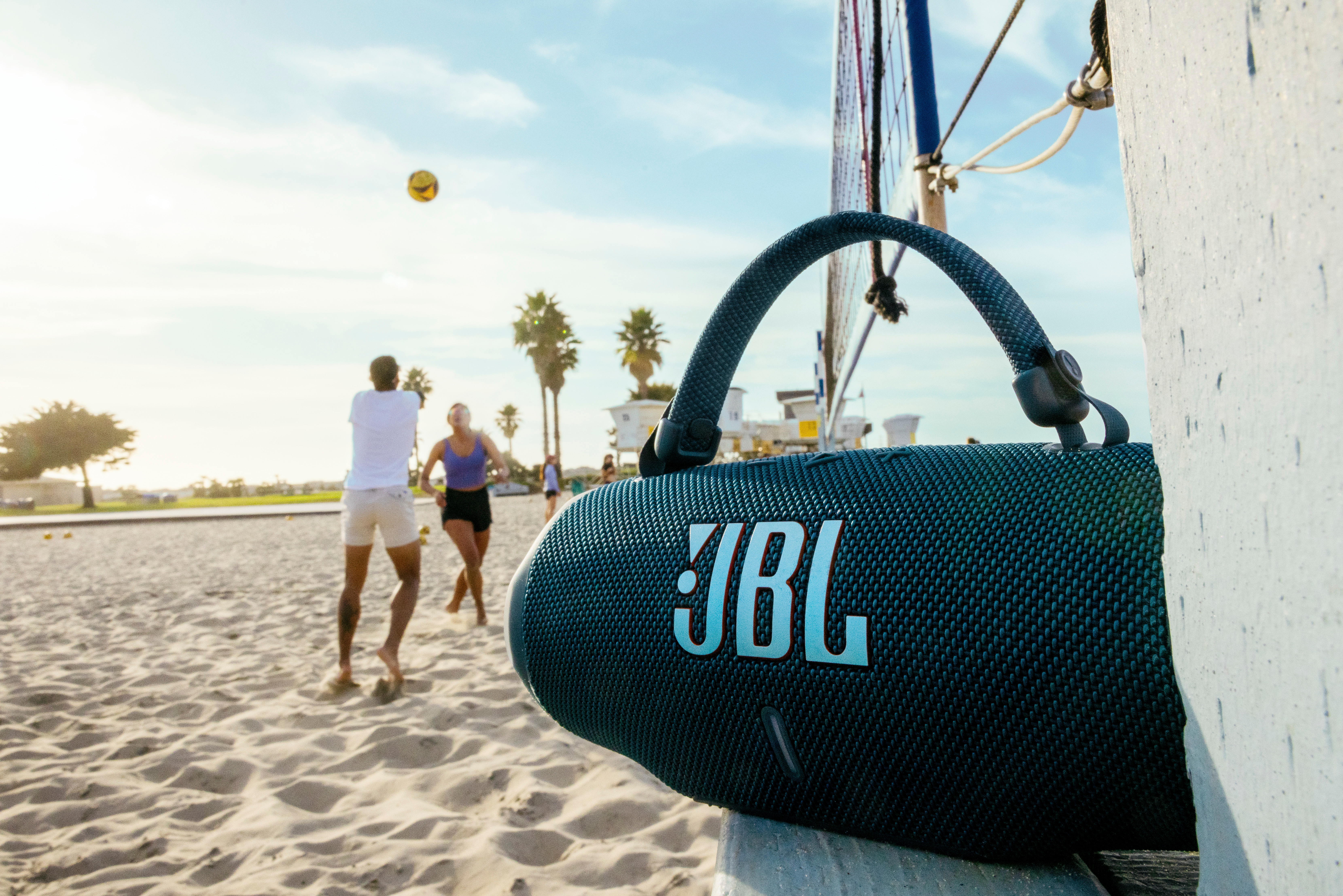 Un altoparlante JBL sulla spiaggia con persone che giocano a pallavolo sullo sfondo.