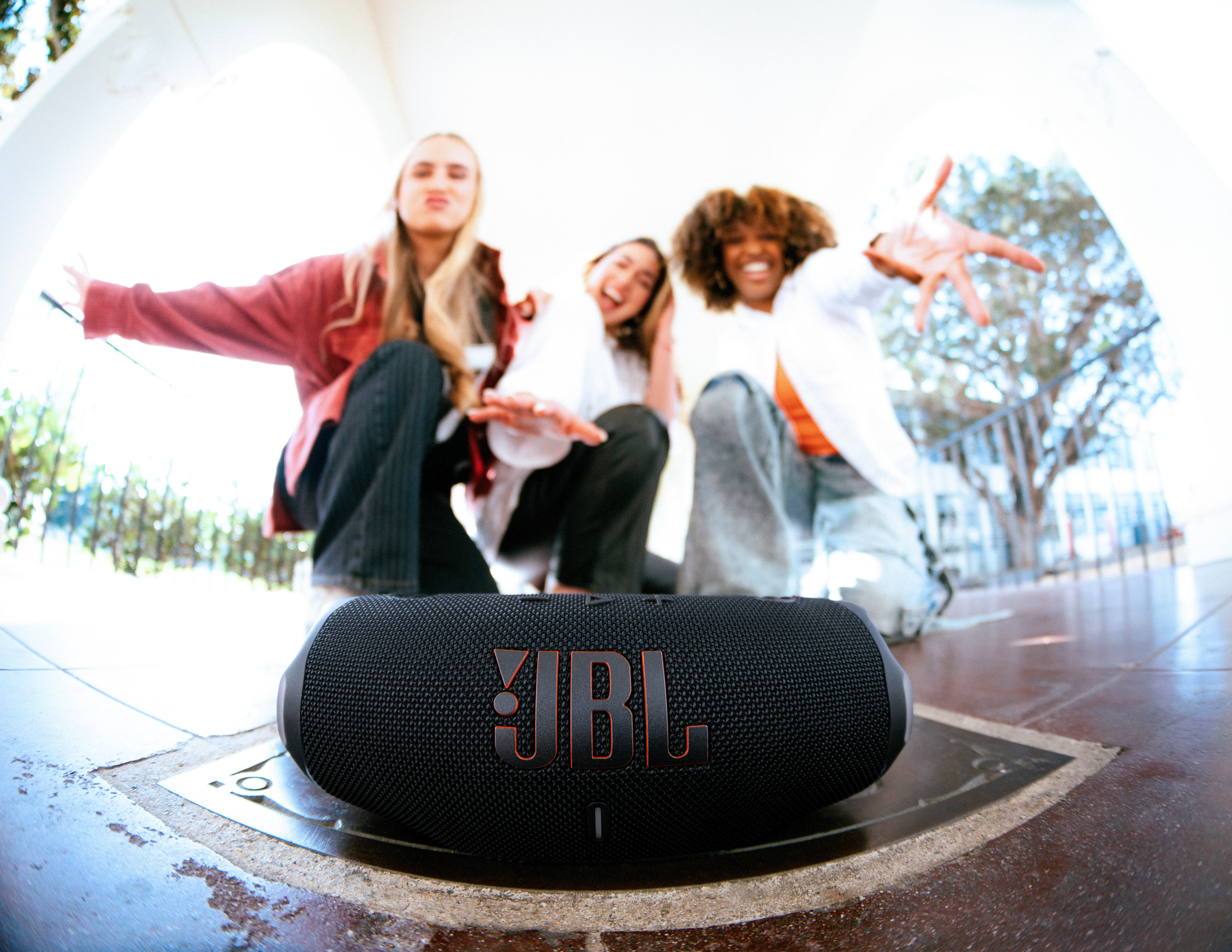 Haut-parleur JBL noir avec logo orange, trois personnes en arrière-plan. La scène est en extérieur.